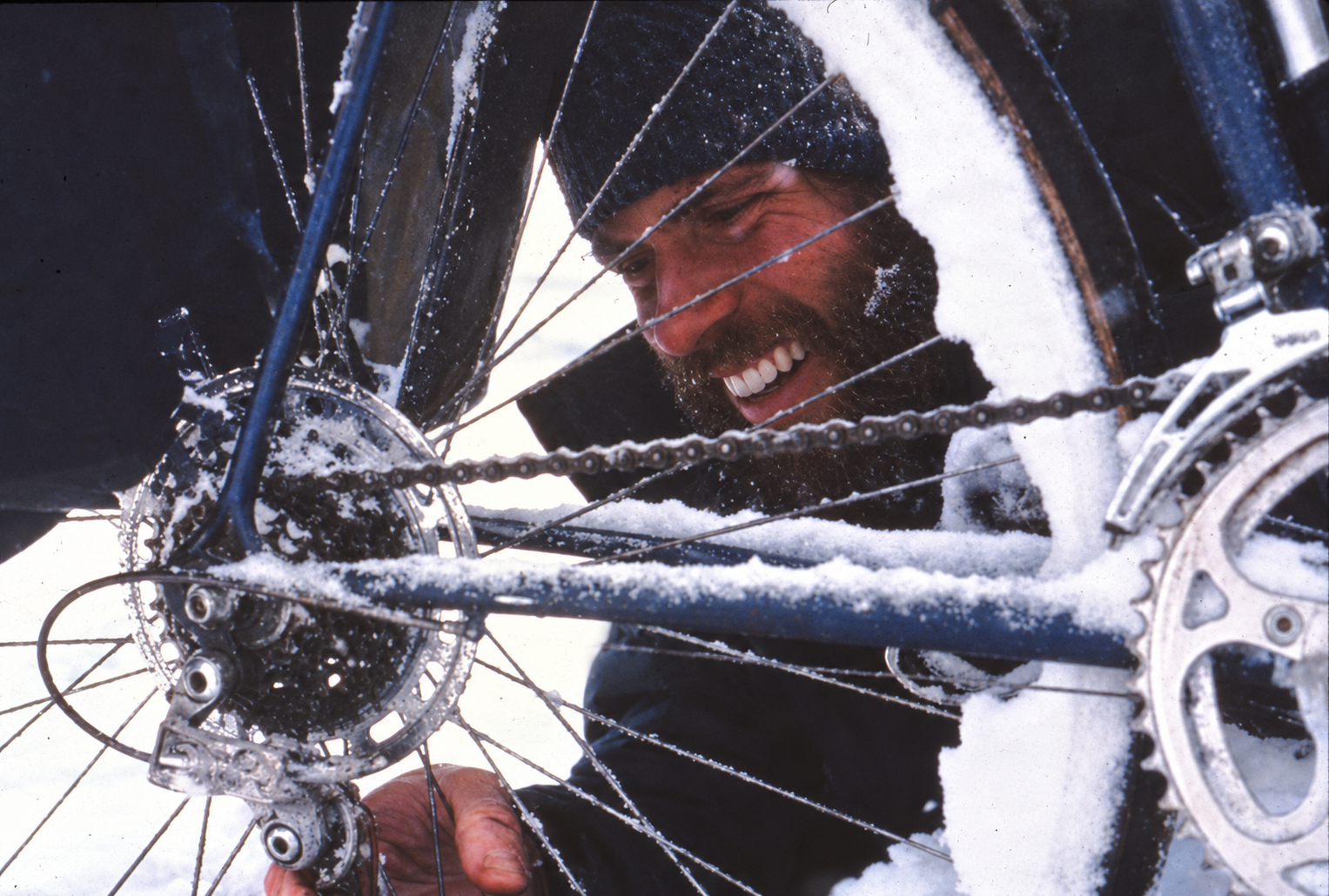 A man with a beard, wearing a black winter hat, is fixing a snow-covered bicycle in a snowy outdoor environment.