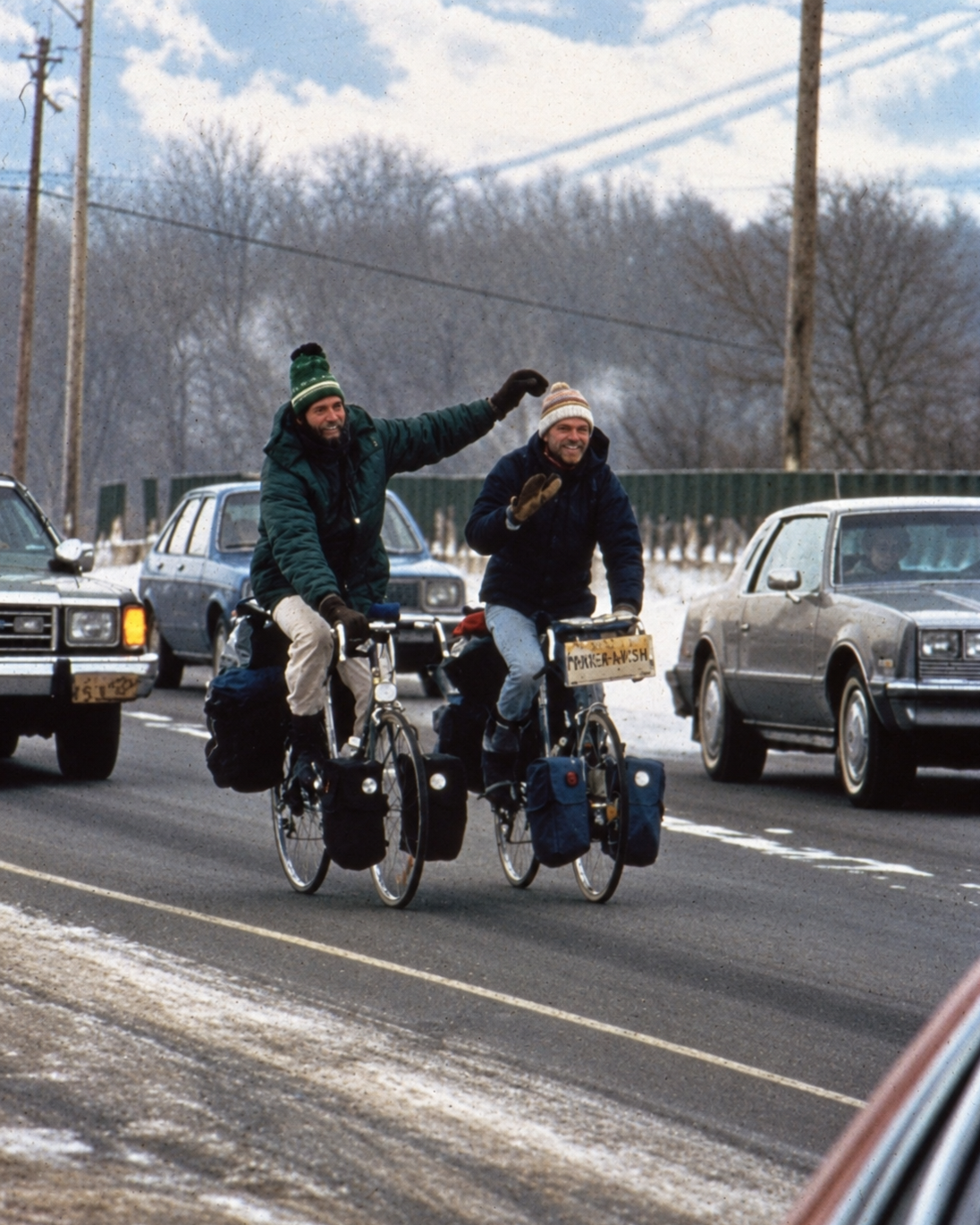 Two men riding tandem bicycles on a snowy road, waving and smiling, with cars parked on the side and trees in the background.