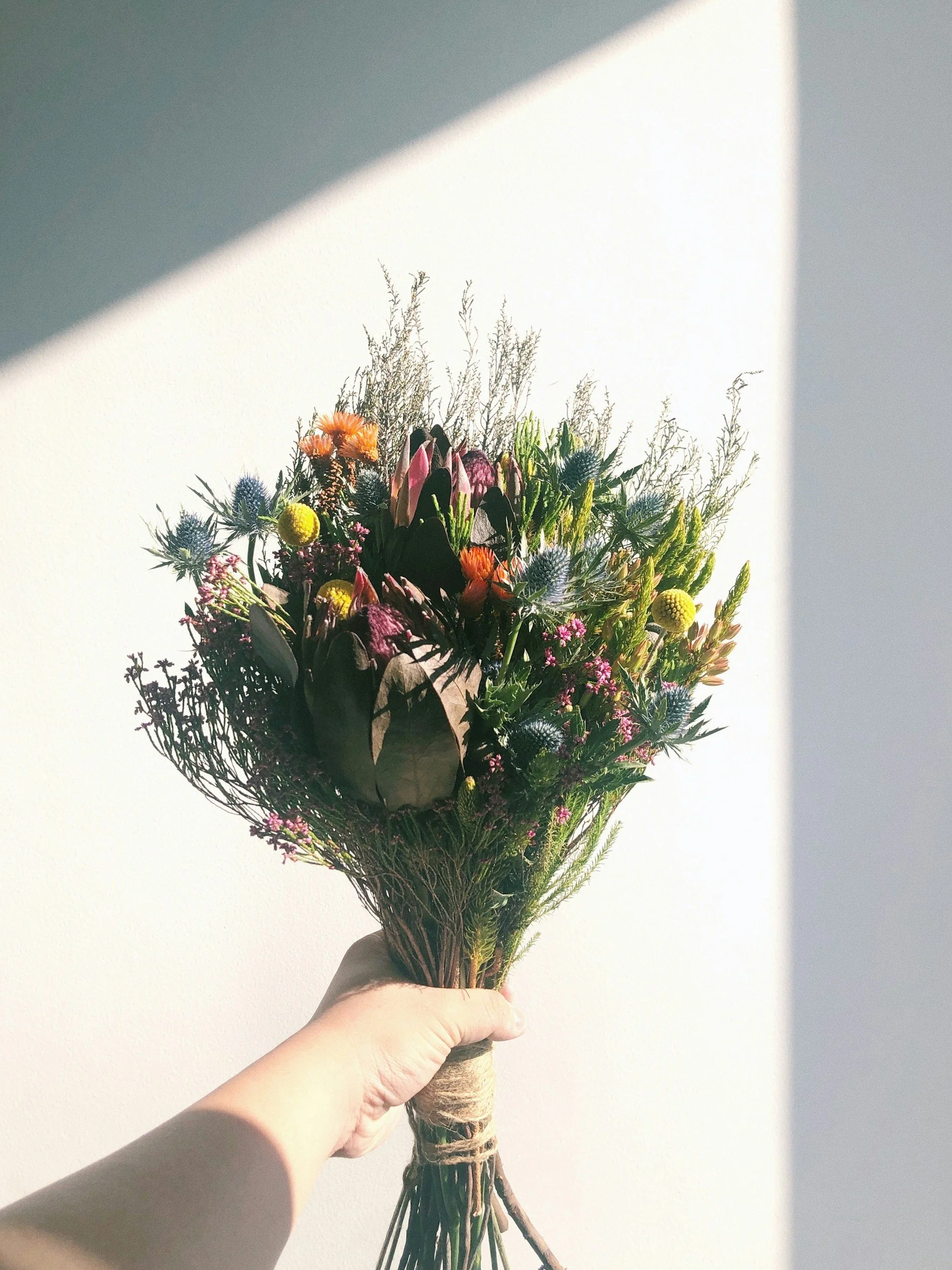 Person holding a colorful flower bouquet against a plain wall with light and shadow.