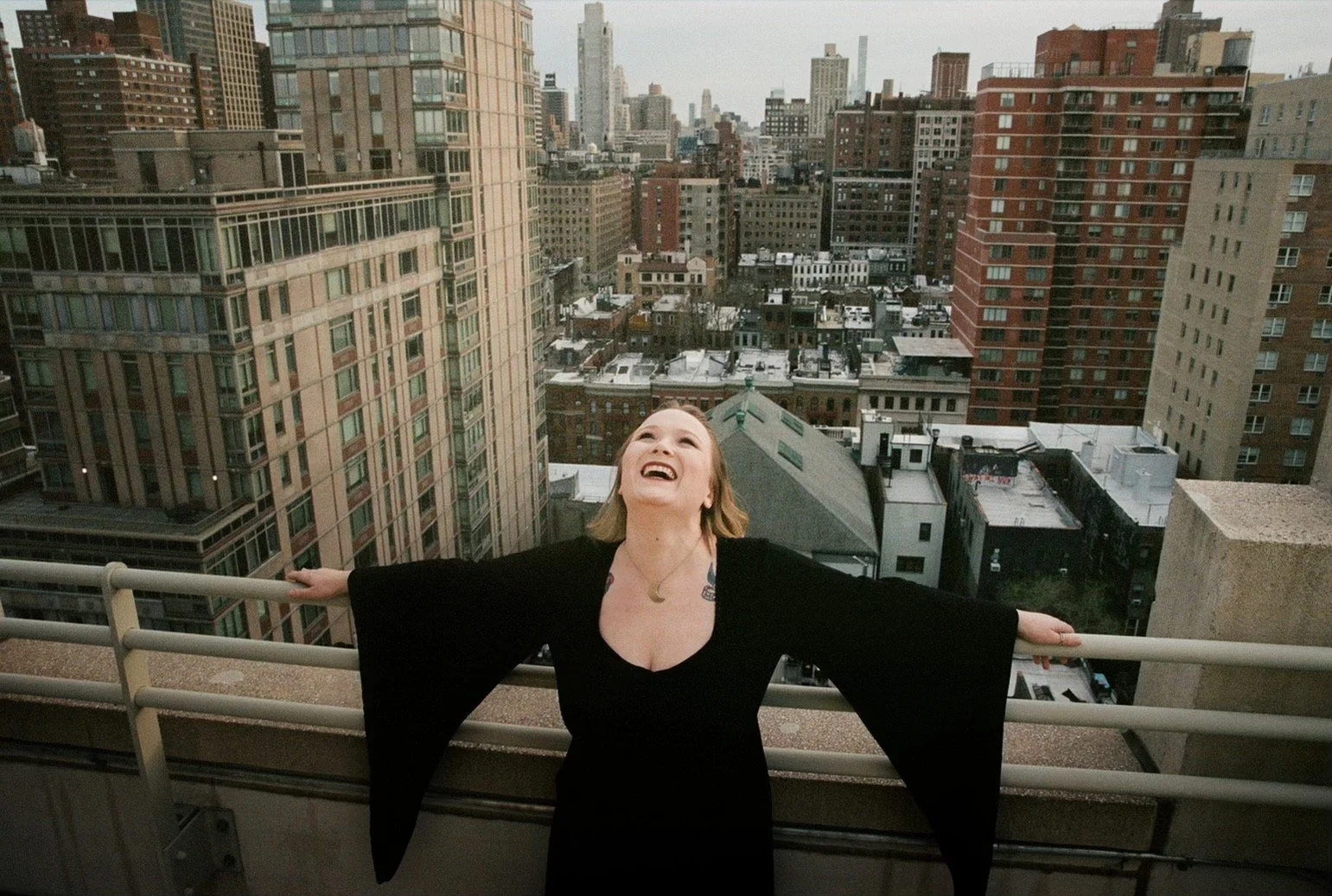 A woman in a black dress standing on a rooftop with an urban cityscape in the background, smiling and looking up with her arms outstretched.