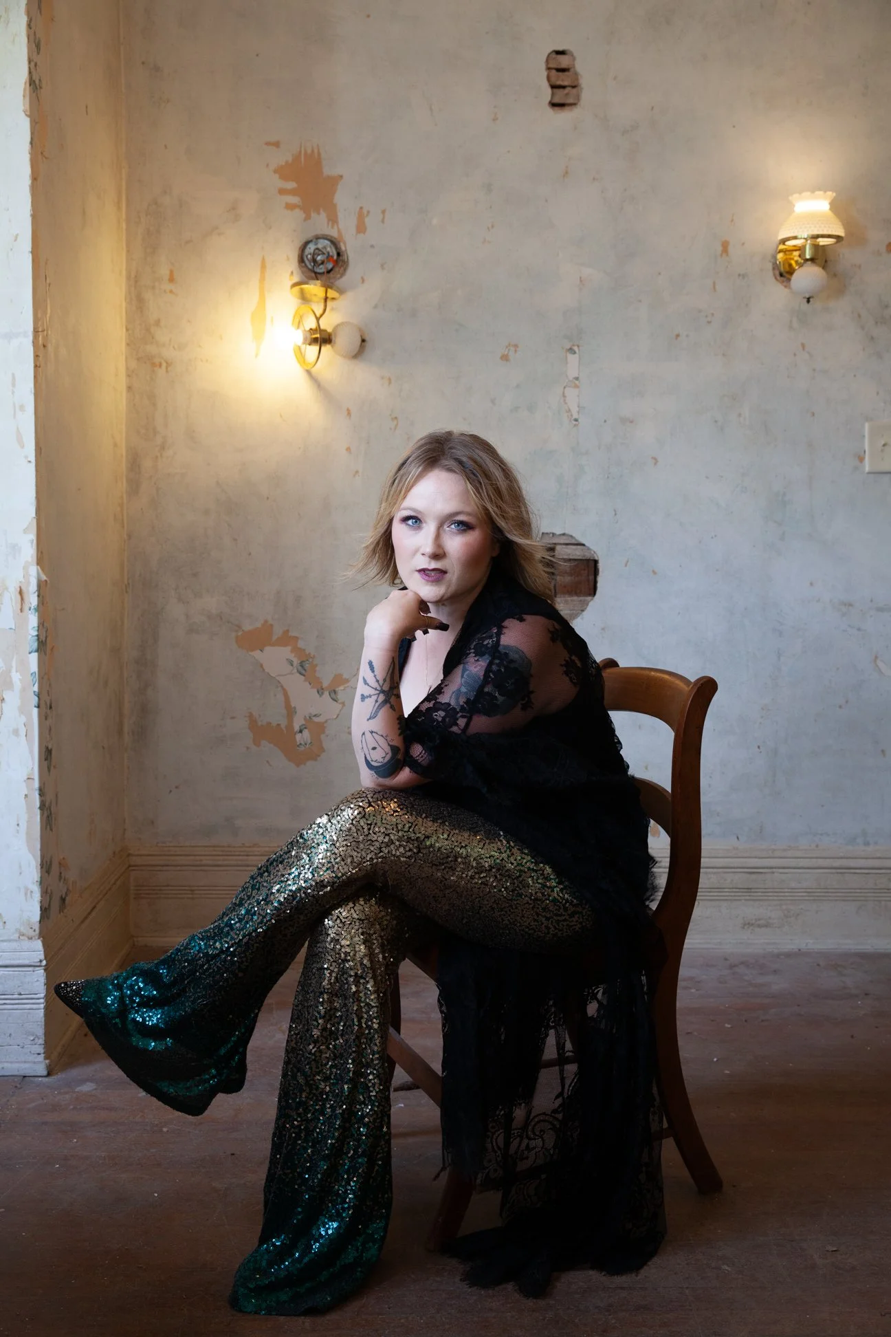A woman with blonde hair wearing a black lace top and gold sequin pants, sitting on a wooden chair in a worn, unfinished room with exposed wall patches and vintage wall sconces lighting.