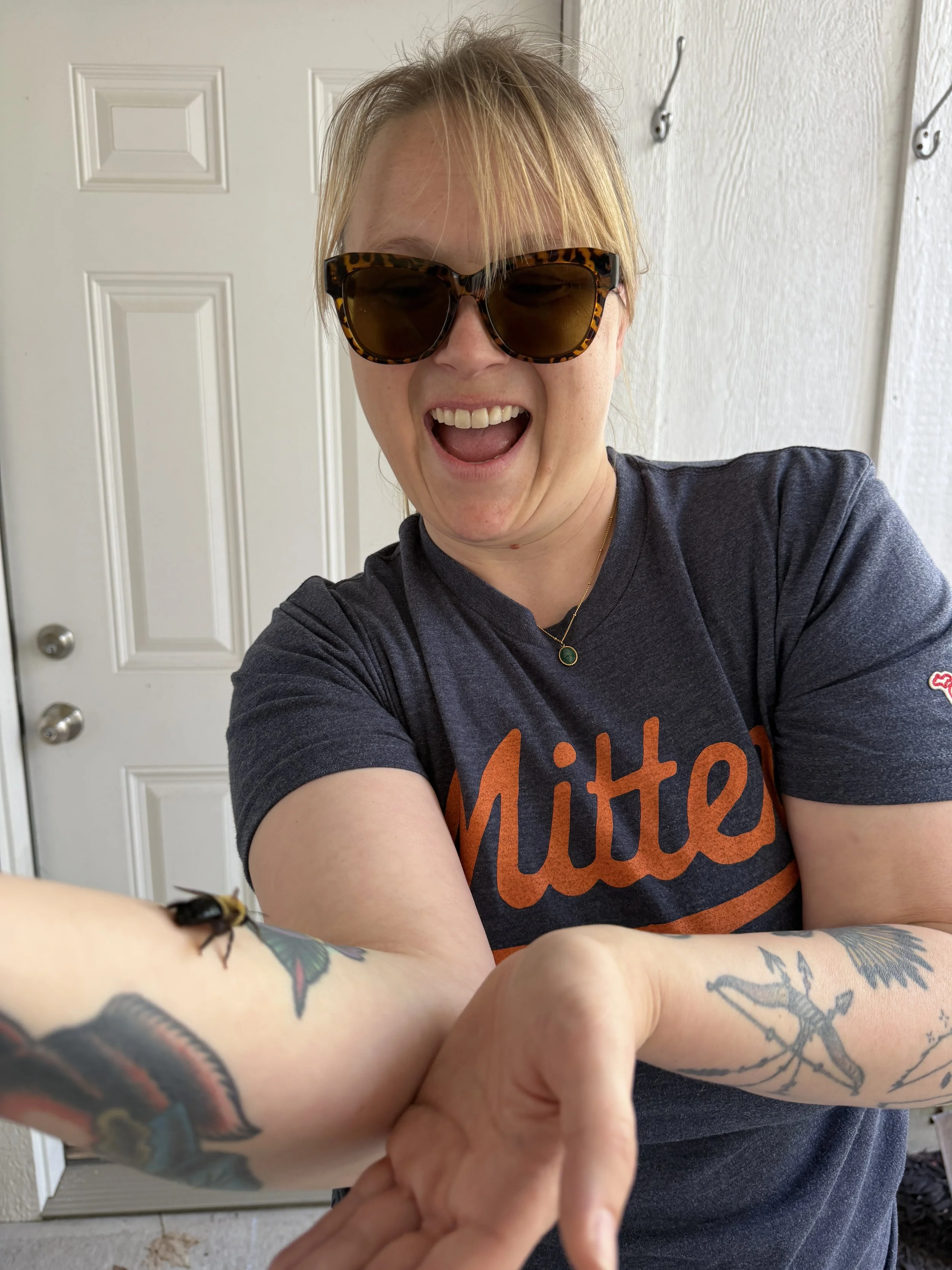 A woman with blonde hair wearing large, tortoise-shell sunglasses smiling widely. She is wearing a dark gray T-shirt with orange text and a necklace with a green pendant. The woman is extending her arms forward, showing tattoos on her forearms, and holding someone else's hand. The background features a white door with decorative paneling and hooks on the wall.