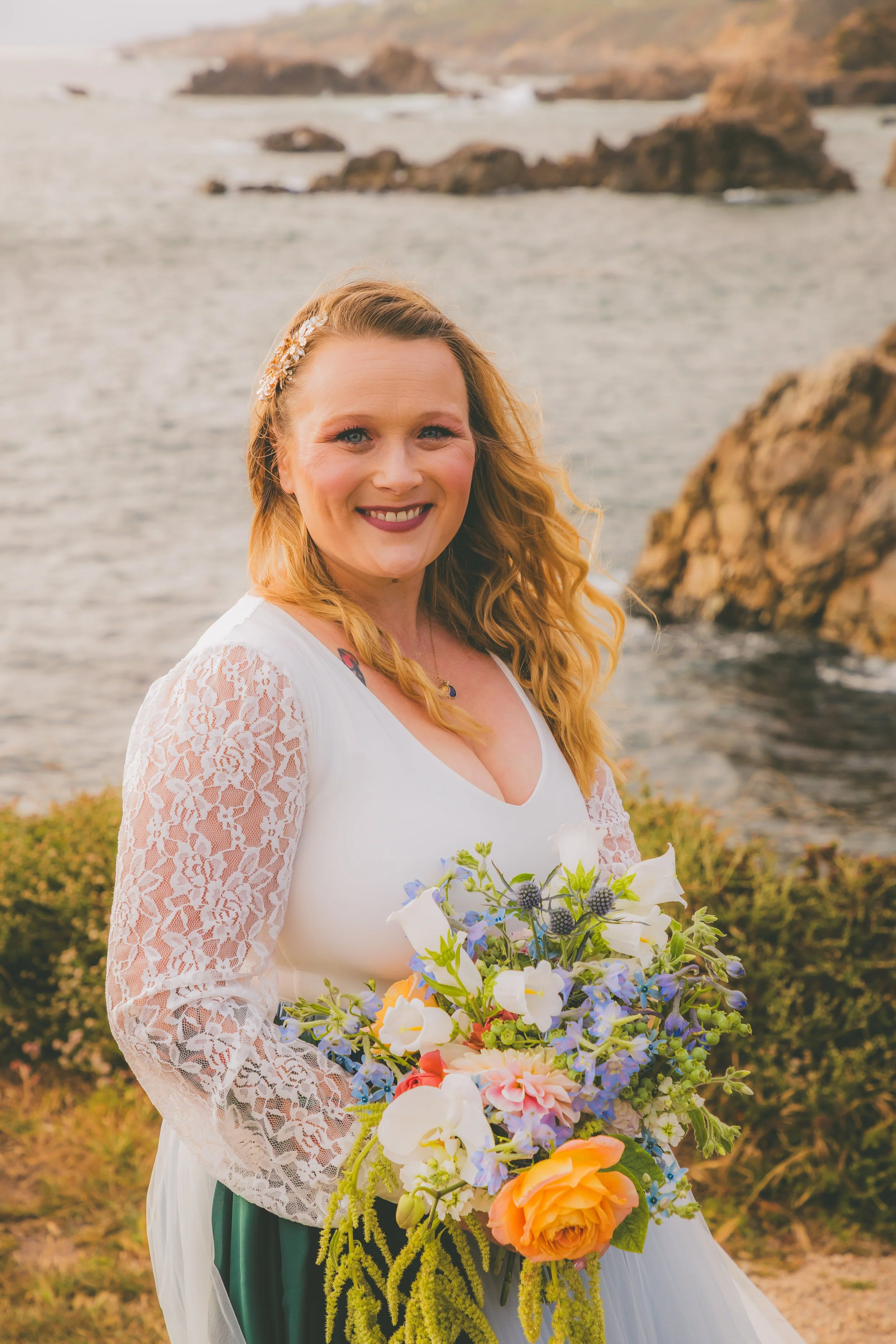 A smiling woman with long, wavy red hair standing outdoors near a rocky shoreline at sunset, holding a large colorful bouquet of flowers.