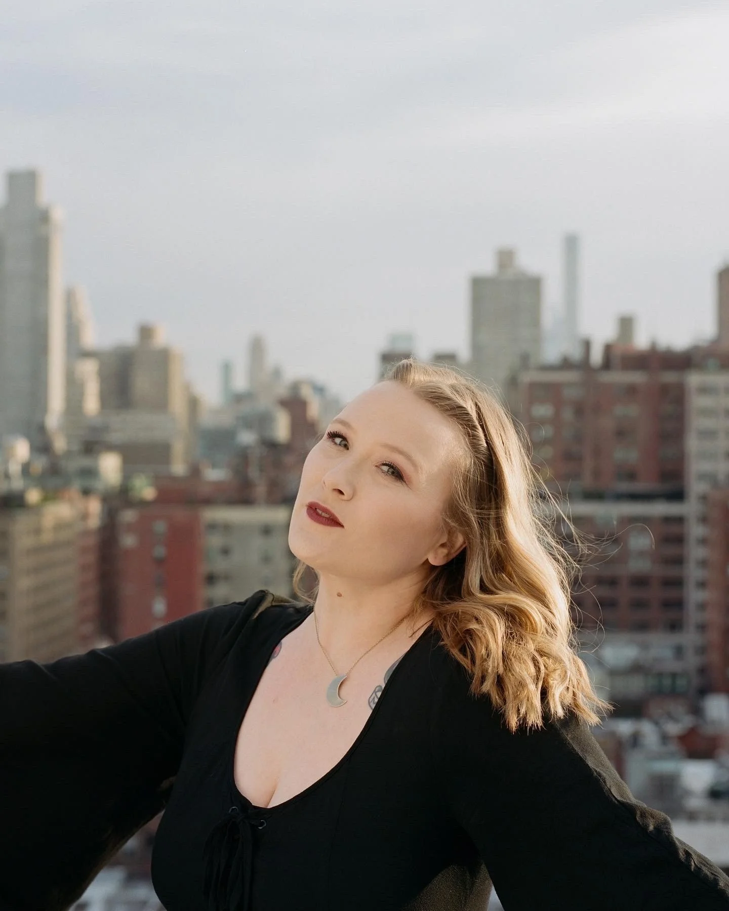 A woman with light skin and wavy blonde hair at a photoshoot on a rooftop with a city skyline in the background.