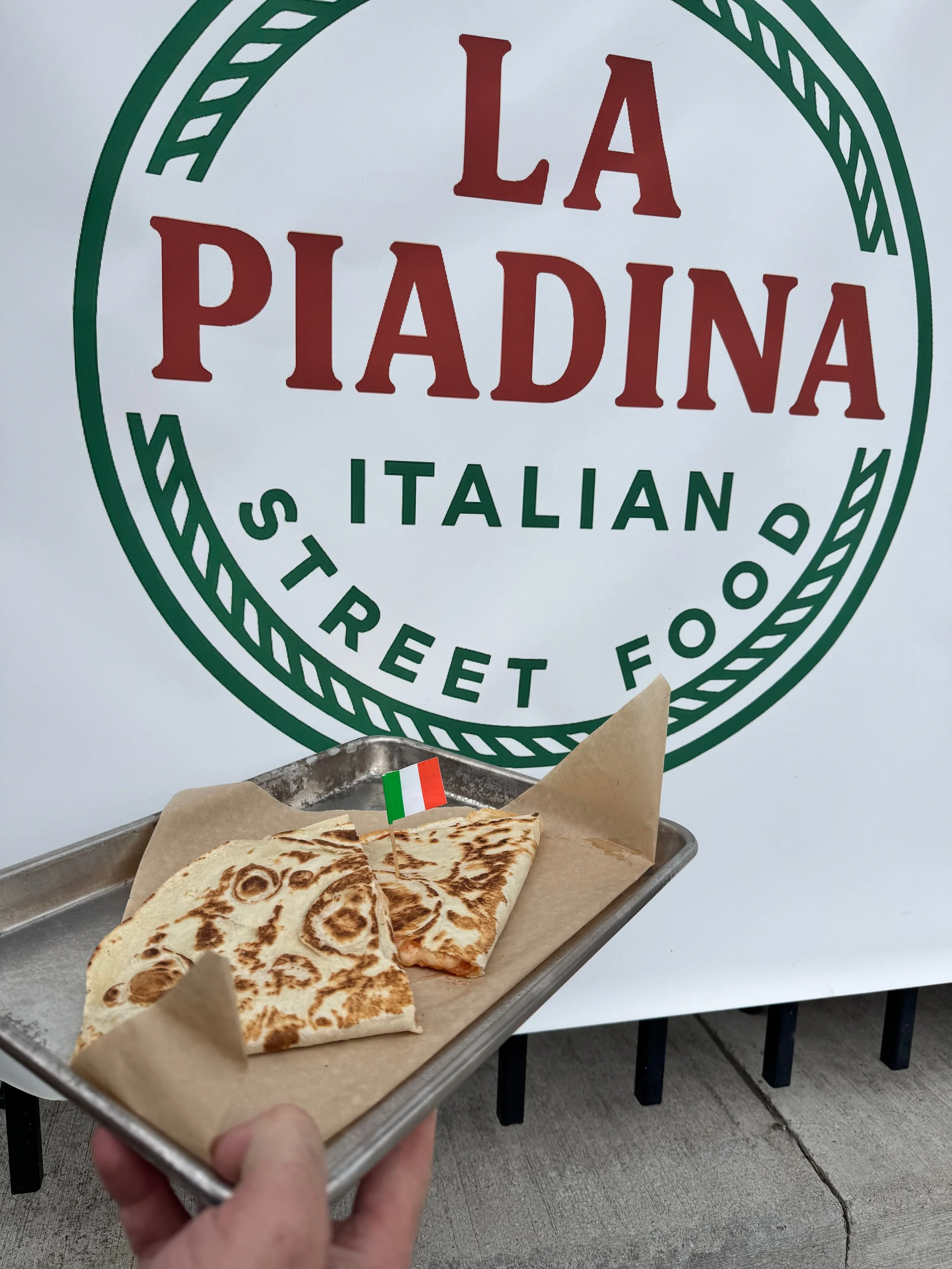 A metal tray with two pieces of grilled flatbread placed on parchment paper, one with a small Italian flag toothpick in it, held in front of a large sign reading 'LA PIADINA - ITALIAN STREET FOOD'.
