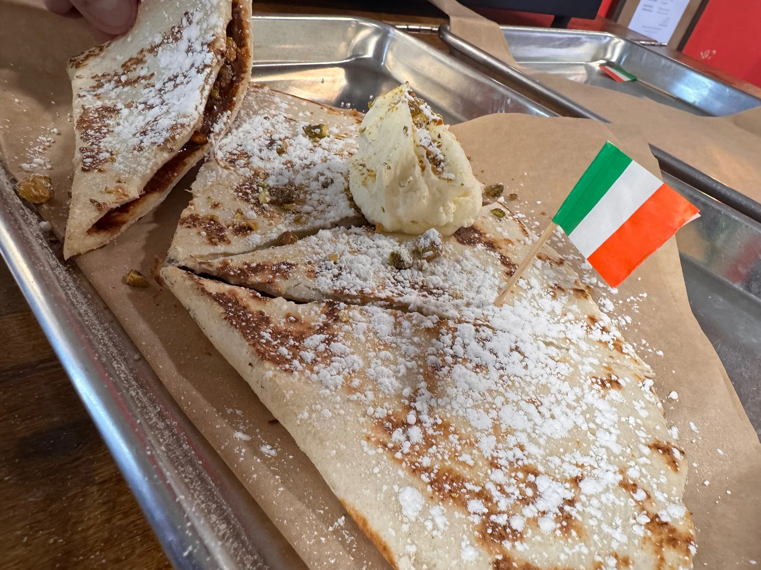 Sliced dessert piadina with white powdered sugar, some green pistachios, a dollop of marscapone cream, and a small Italian flag on a metal tray.