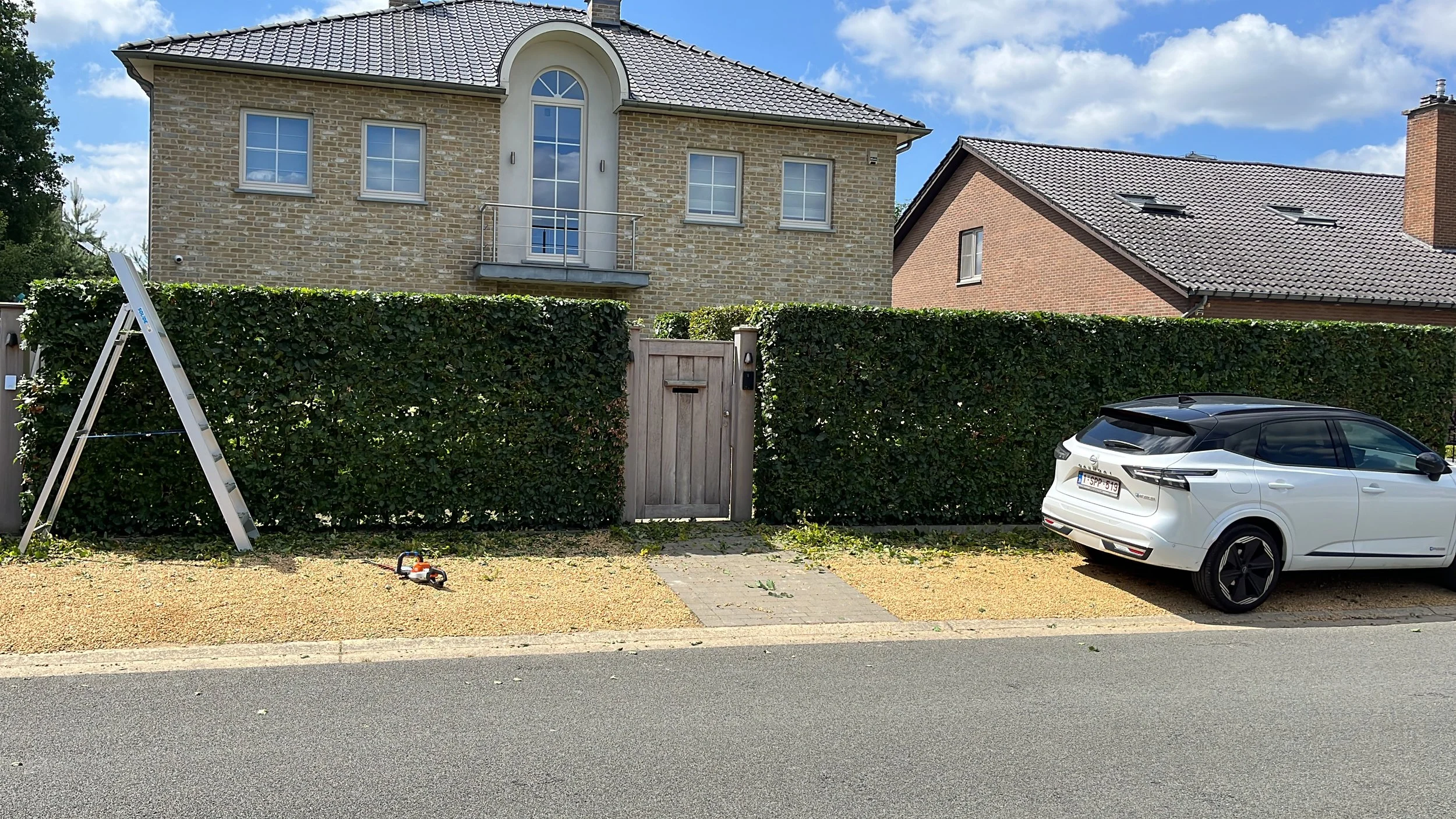 A residential house with a brick exterior, white framed windows, and a small balcony, surrounded by a tall hedge and a wooden gate. In the front yard, there is a white electric car parked on a gravel surface, and a small toy car lying on the ground n
