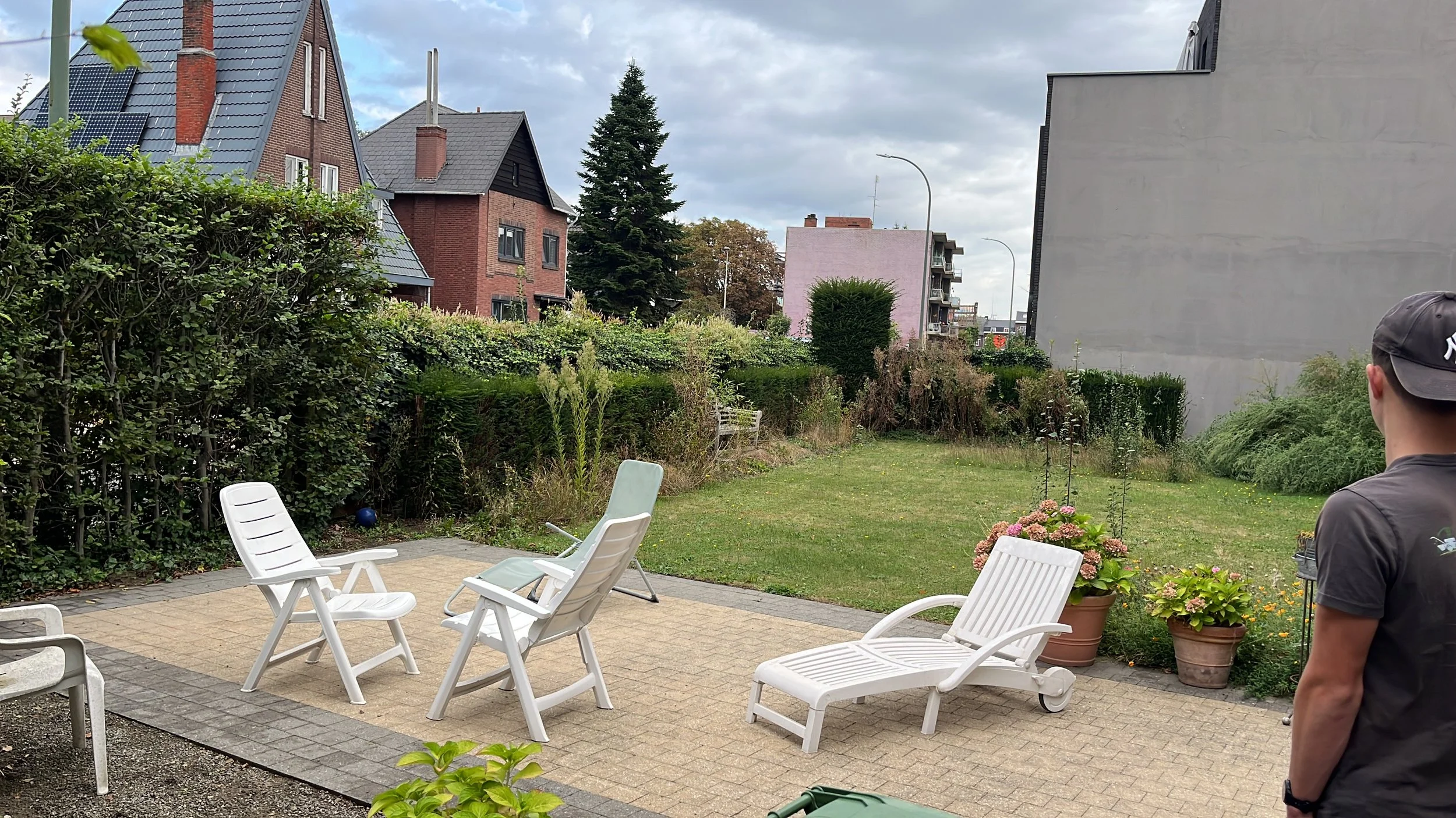 Backyard patio with white outdoor chairs, potted plants, a man wearing a gray cap and t-shirt, surrounded by a hedge and neighboring houses