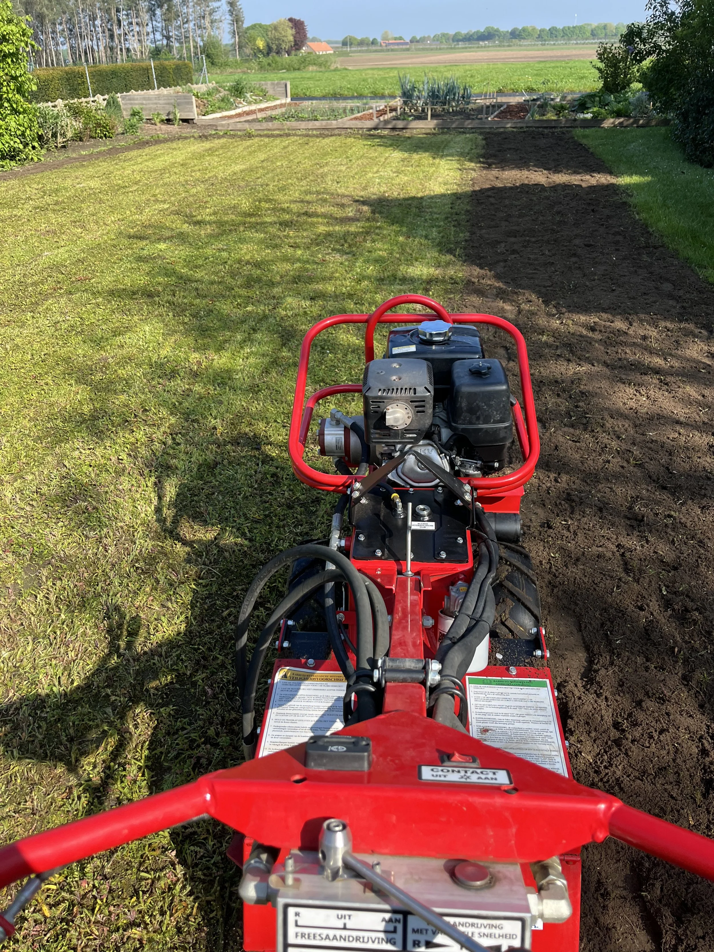 A red rotary tiller machine operating on a lawn and garden bed, with green fields and farm buildings in the background.