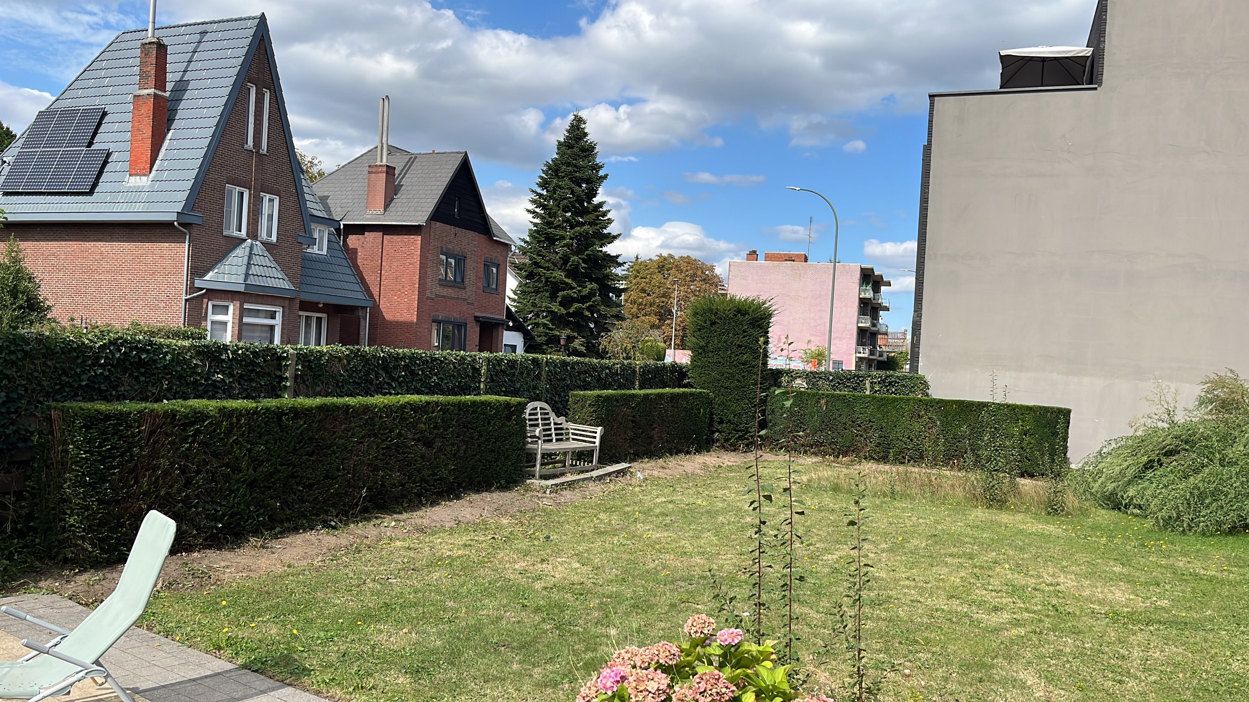 A backyard garden with a bench, a lawn, and neatly trimmed hedges, surrounded by multi-story residential buildings under a partly cloudy sky.