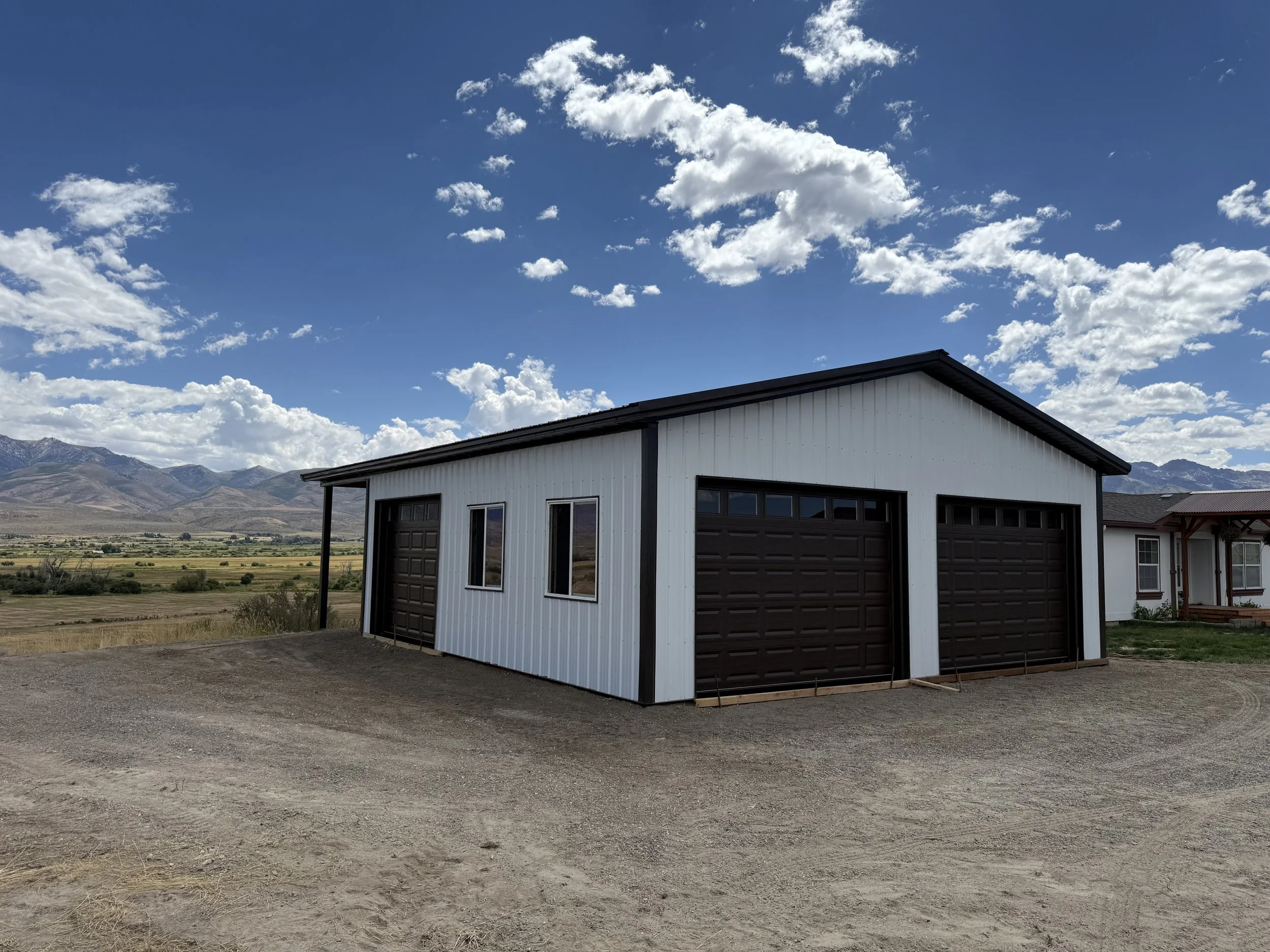 A white metal garage with two brown garage doors and two windows, set on a dirt driveway in a rural area with mountains and a blue sky with clouds in the background.
