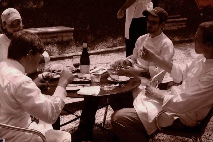 Four people dining outdoors at a round table, with glasses, plates, and a bottle of wine, engaging in conversation.