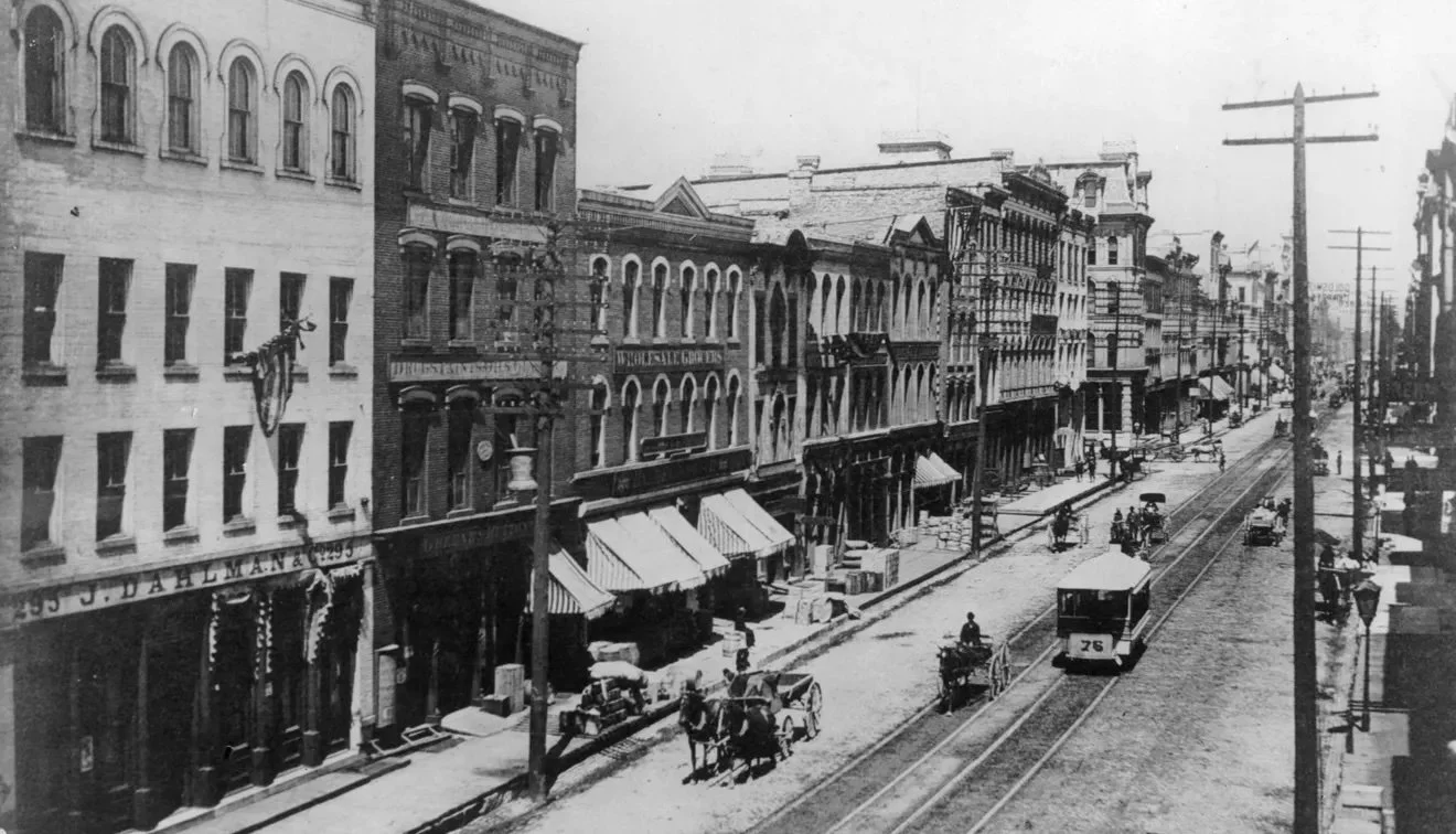 Historical black and white photo of a city street with buildings, streetcars on tracks, pedestrians, horse-drawn carriages, and telephone poles.