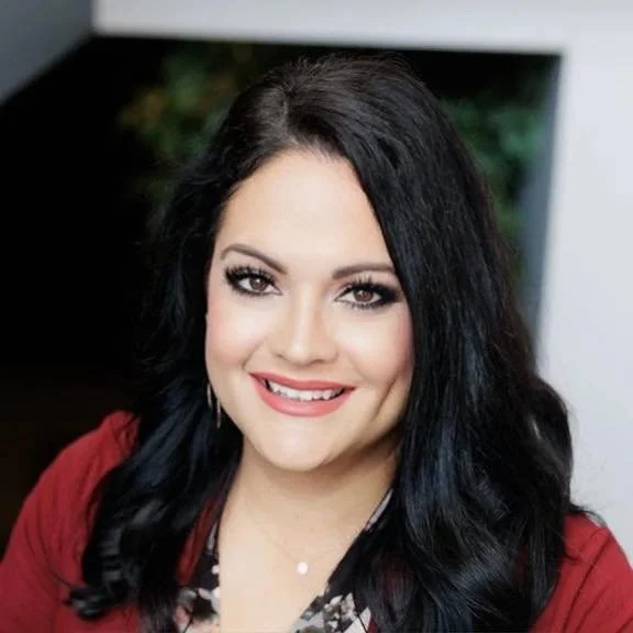 A woman with long black hair smiling at the camera, wearing a red top and makeup, in an indoor setting.