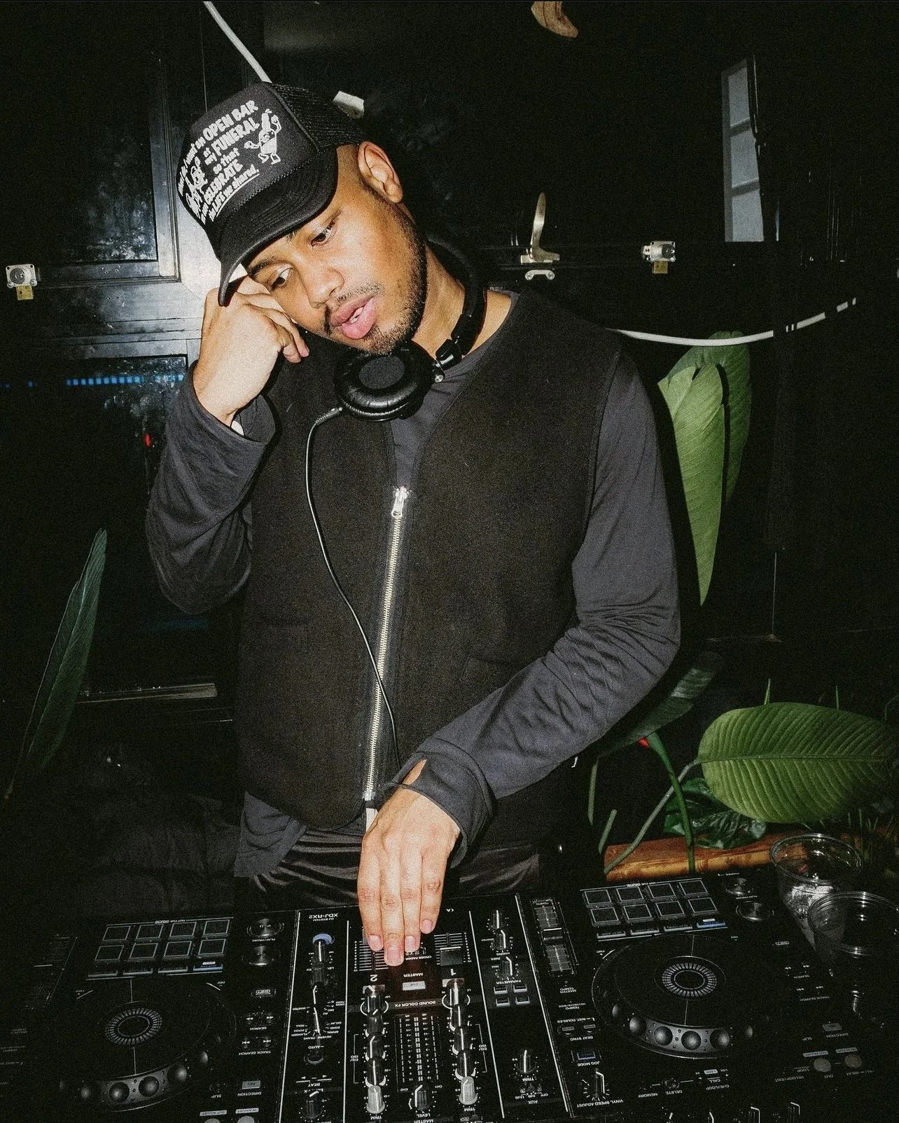 A DJ with headphones around his neck, wearing a black vest and a cap, standing behind a DJ console with turntables and controls, in a dark indoor setting with green plants in the background.