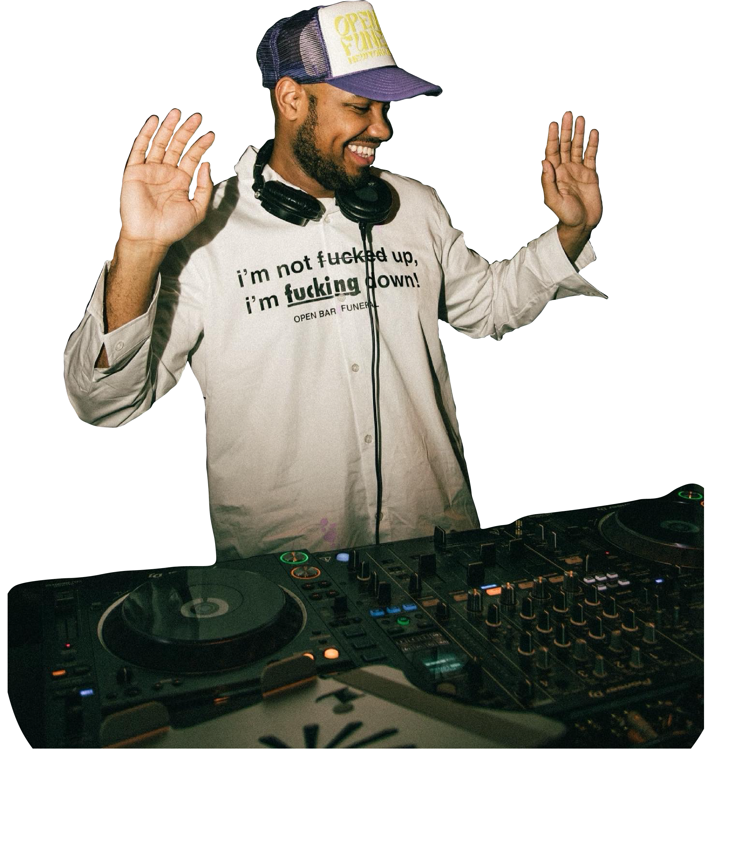 A male DJ wearing a white shirt with a humorous print, a trucker hat, and headphones around his neck is dancing or DJing behind a DJ console. He has a broad smile, eyes closed, and hands raised.