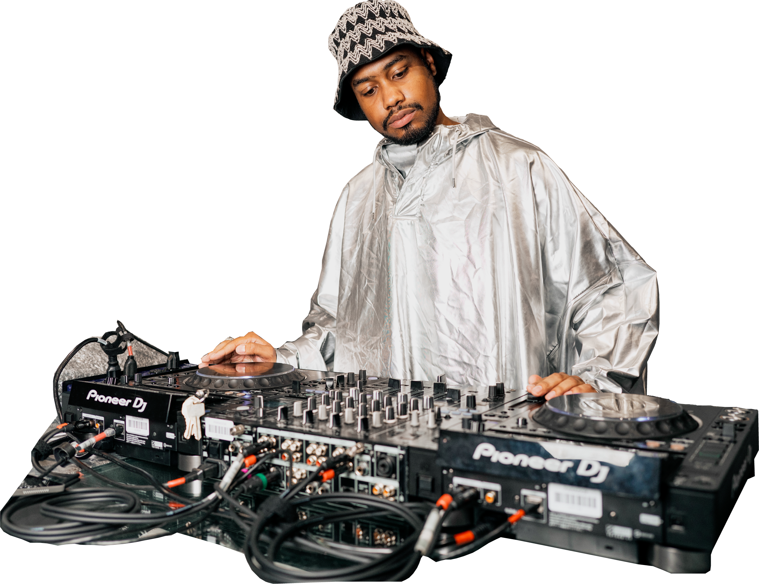 A DJ wearing a silver jacket and patterned hat, standing behind DJ equipment with turntables and a mixer, operating the controls.