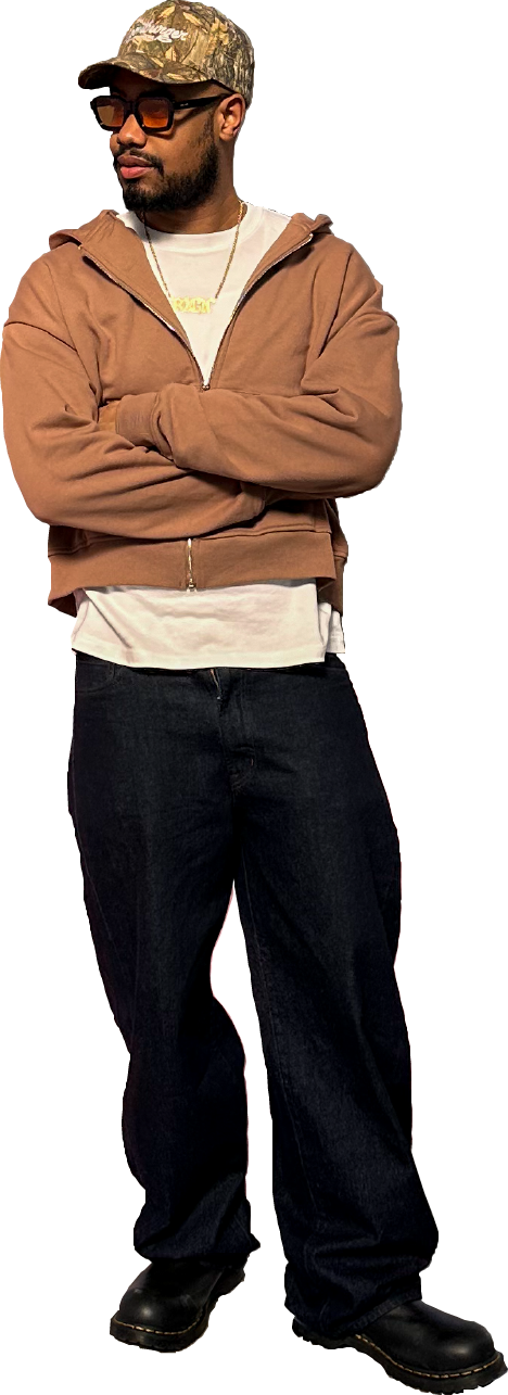 A man standing with his arms crossed, wearing a camouflage baseball cap, dark sunglasses, a tan zip-up hoodie, a white t-shirt underneath, black jeans, and black boots.