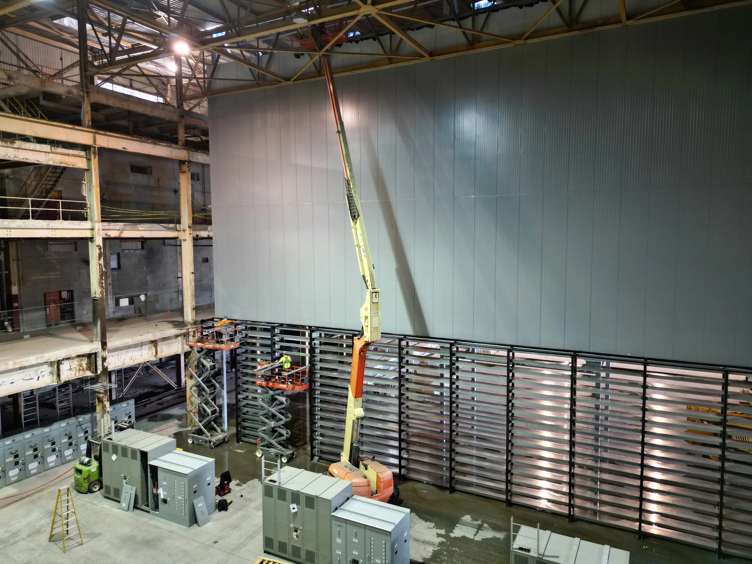 Construction inside a factory with machinery, scaffolding, and a large gray wall being assembled.