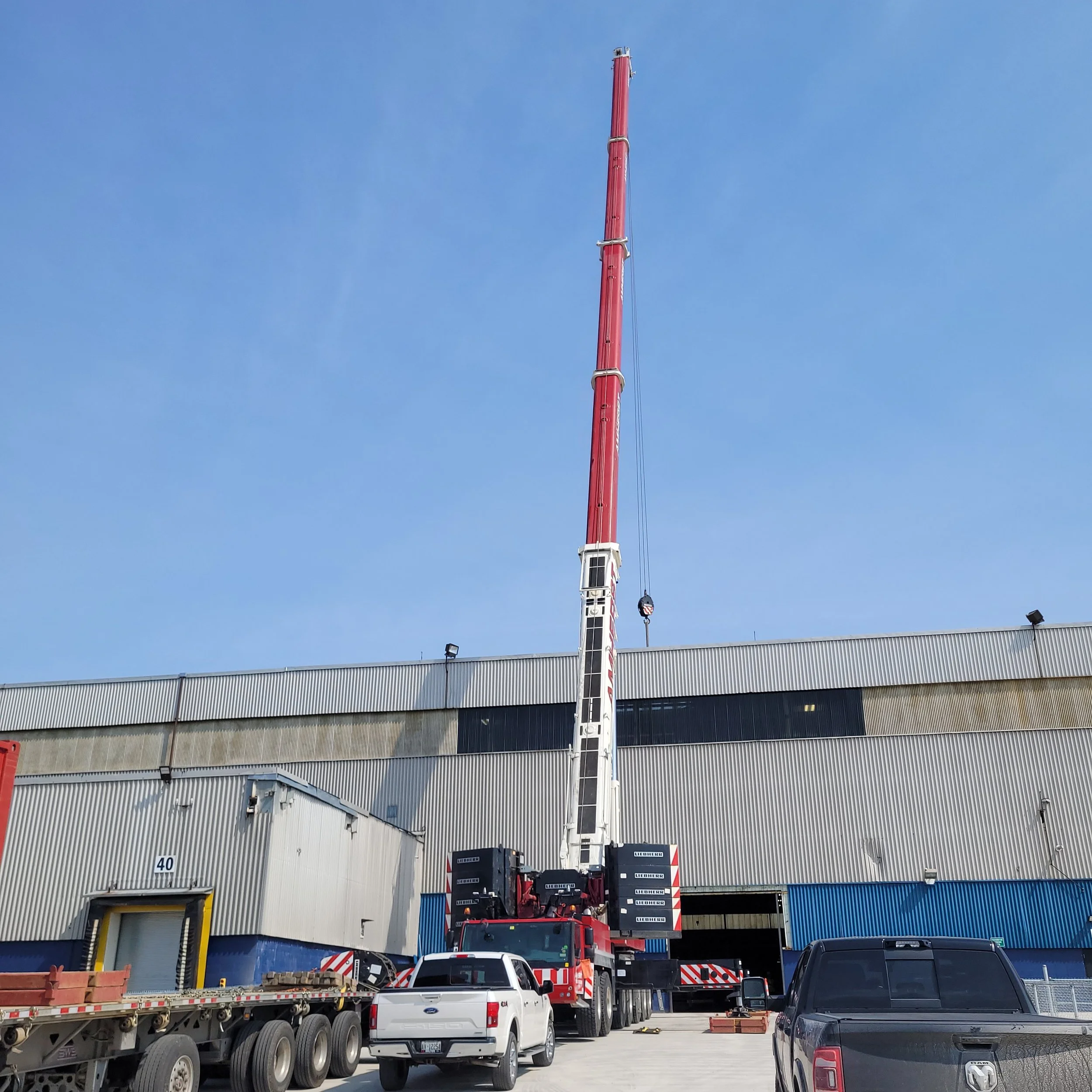Large red and white mobile crane with extended jib stabilizing in front of an industrial warehouse, with trucks and vehicles parked nearby on a sunny day.