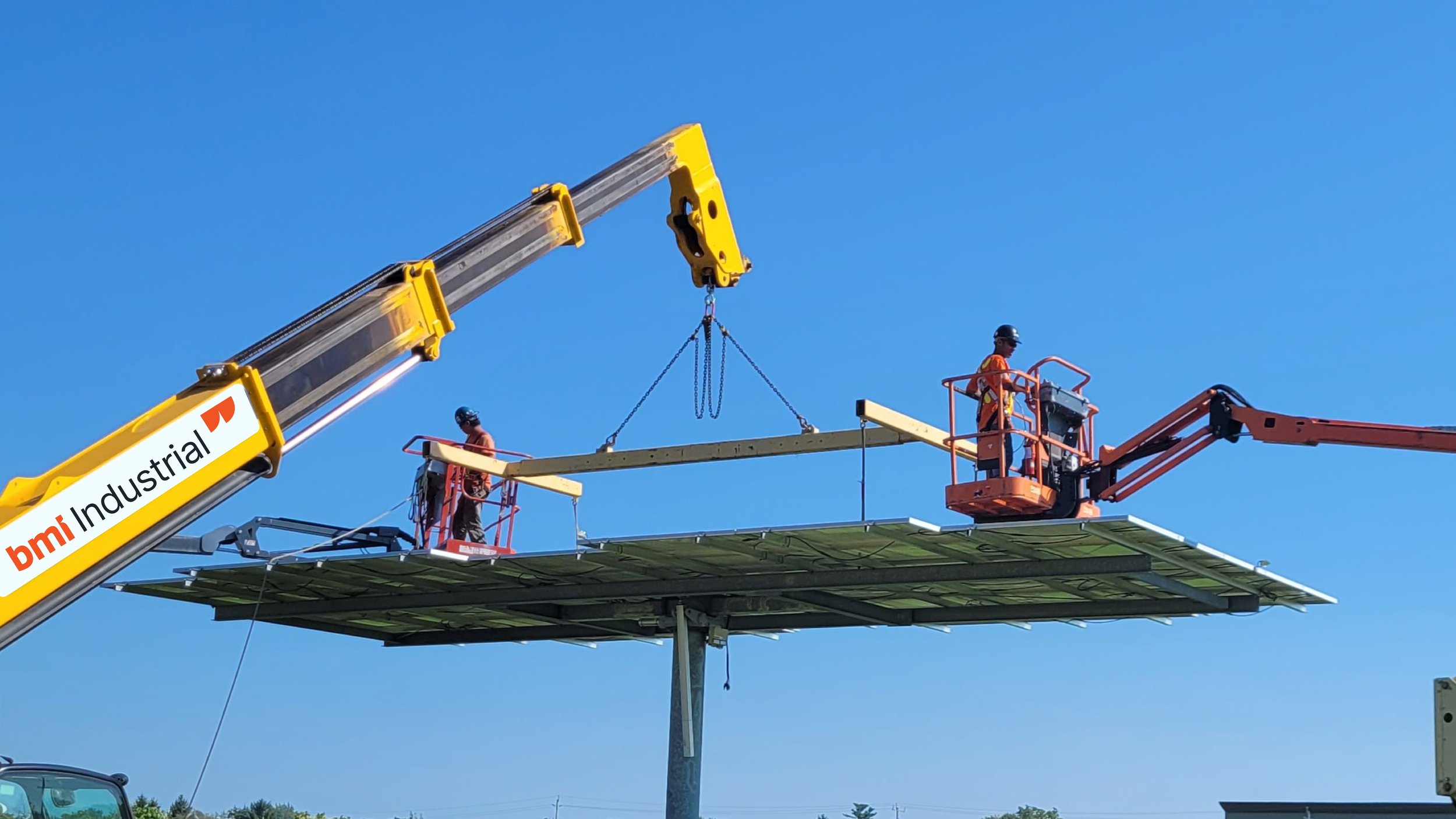 Construction workers using a crane and lift to assemble solar panels on a platform, under a clear blue sky.