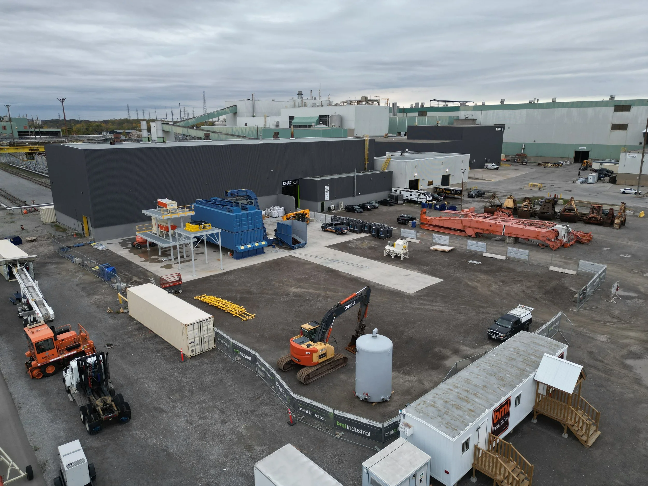 Industrial construction site with heavy machinery, storage containers, construction vehicles, and a large industrial building under cloudy skies.