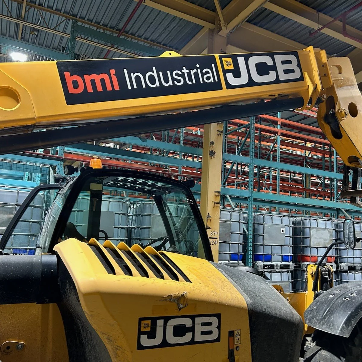 Yellow industrial JCB machine with a black and white JCB logo and a black and red bmj Industrial sticker, inside a warehouse with metal shelving and stored tanks.
