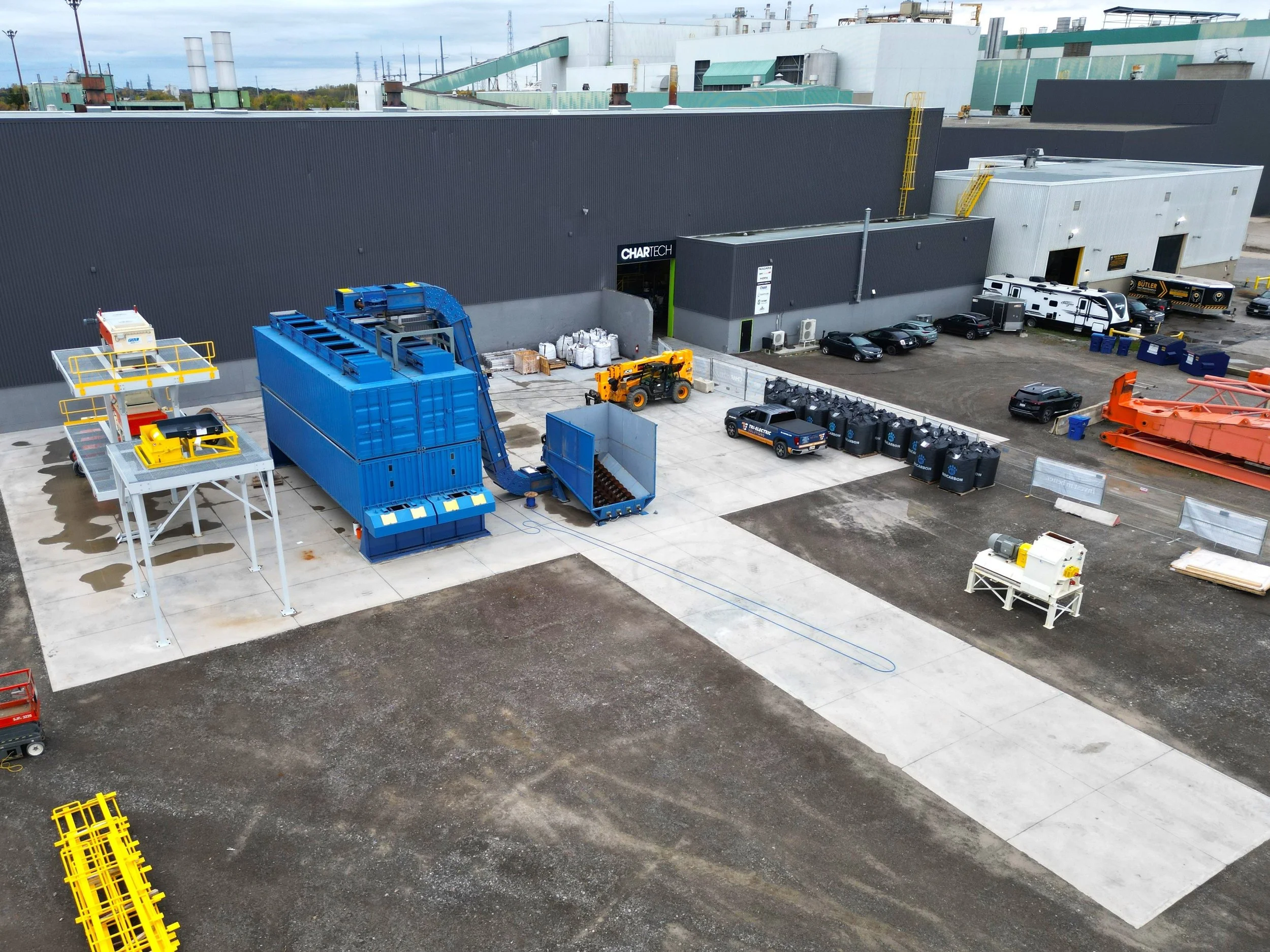 Industrial outdoor scene with large blue machine, trucks, cars, and construction equipment near a gray building with the sign 'CHARTECH'.
