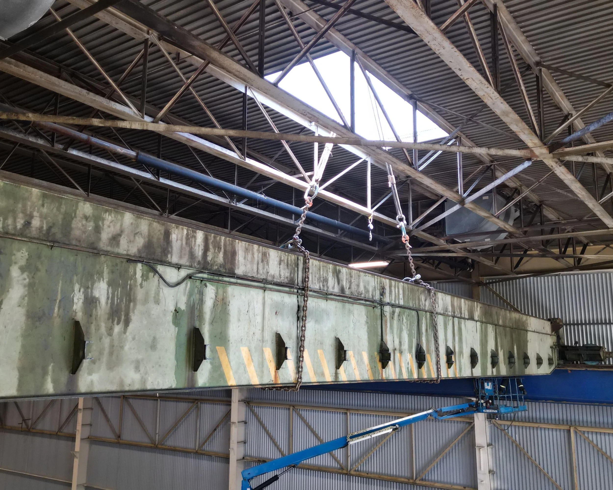 Large industrial or cargo container suspended in a warehouse with metal roof and beams, supported by a blue lifting machine.