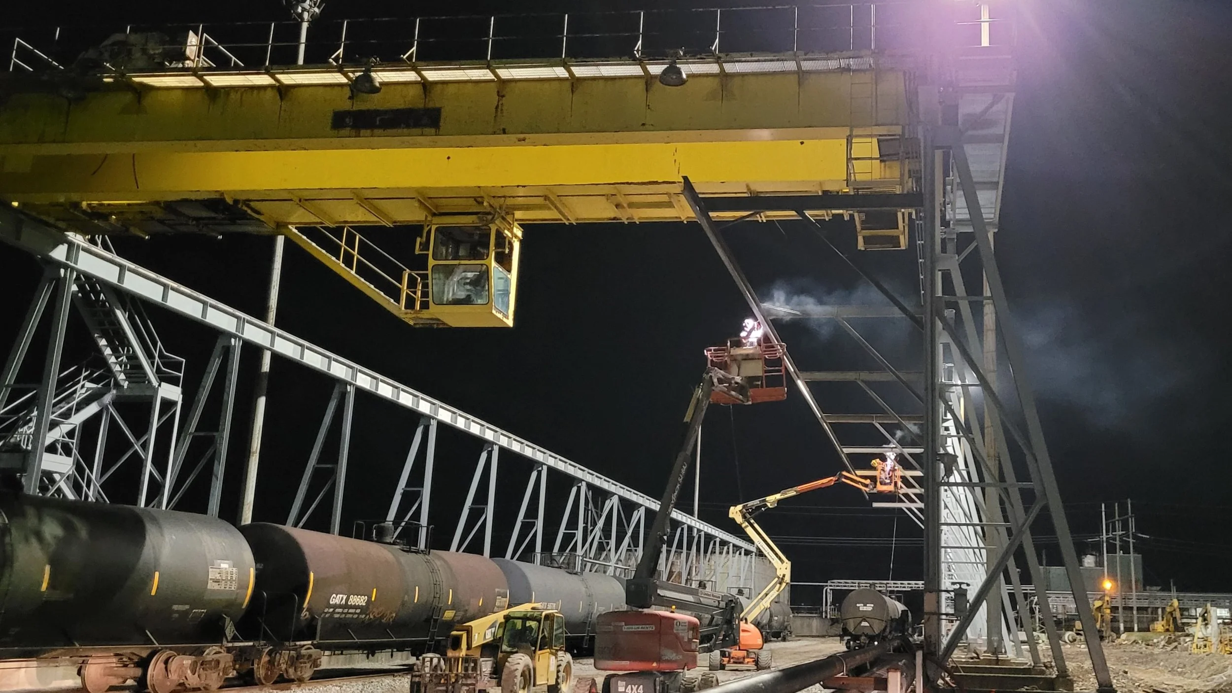 Night scene of construction work on a railway with workers welding on a large metal structure. Multiple cranes and boom lifts are used to access high sections of the structure. Railway tracks, a black oil tanker train car, and construction equipment 