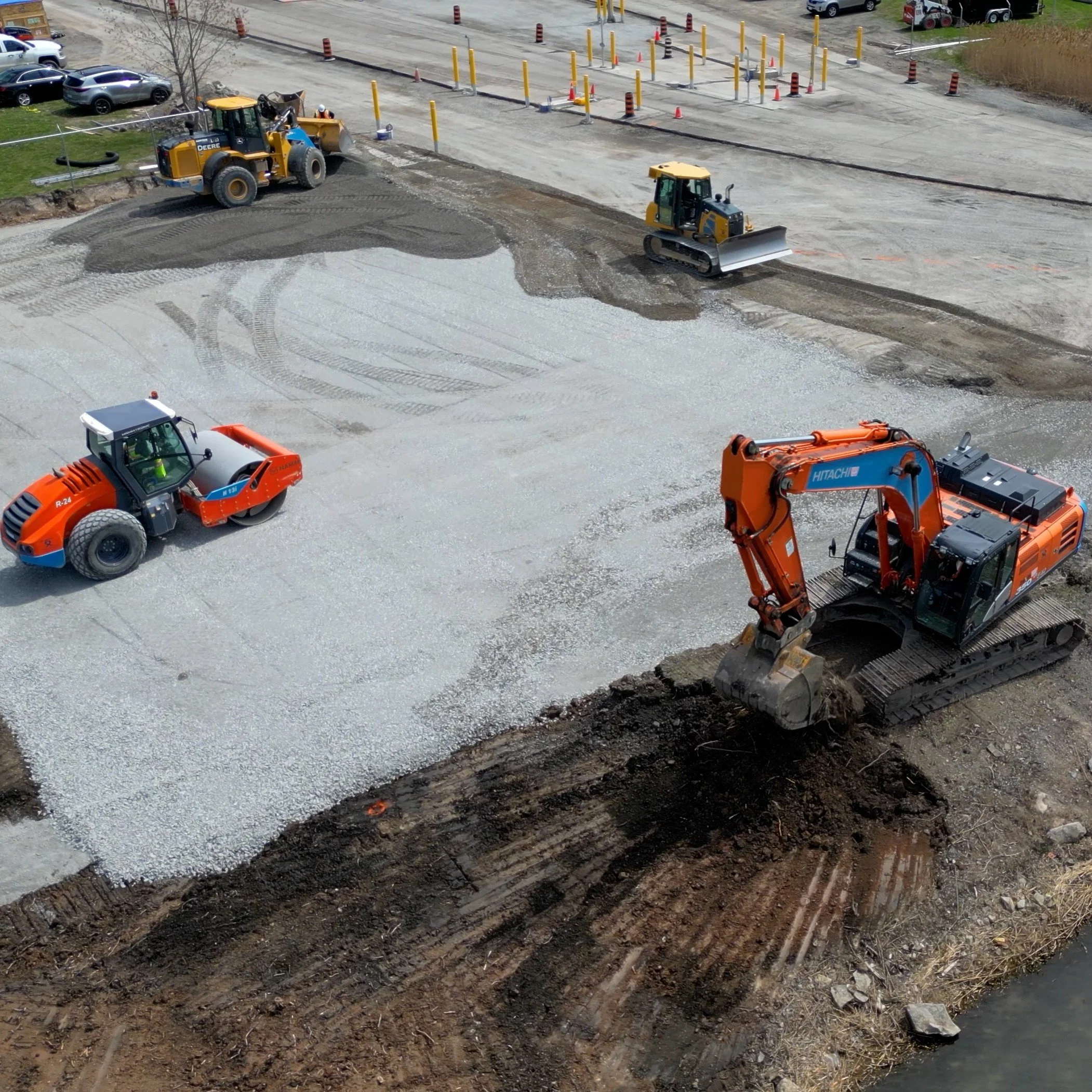 Construction site with heavy machinery, including a large orange excavator and a steamroller, working on leveling and preparing the ground for paving.