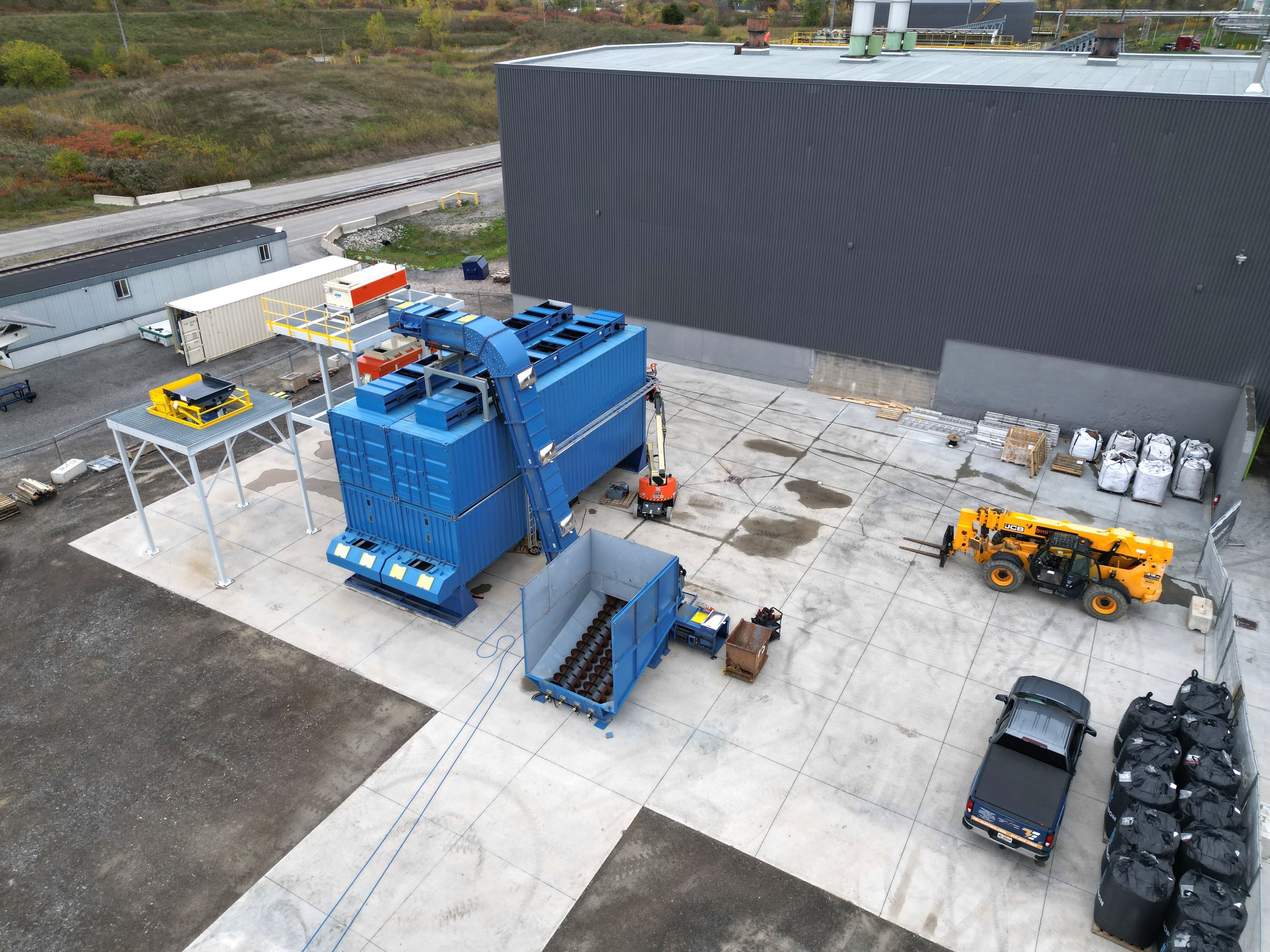 An industrial outdoor area with a large gray building, a yellow forklift, several large black bags on pallets, a blue container with a conveyor system, a car, and various equipment and materials, on a concrete surface surrounded by a fenced area.