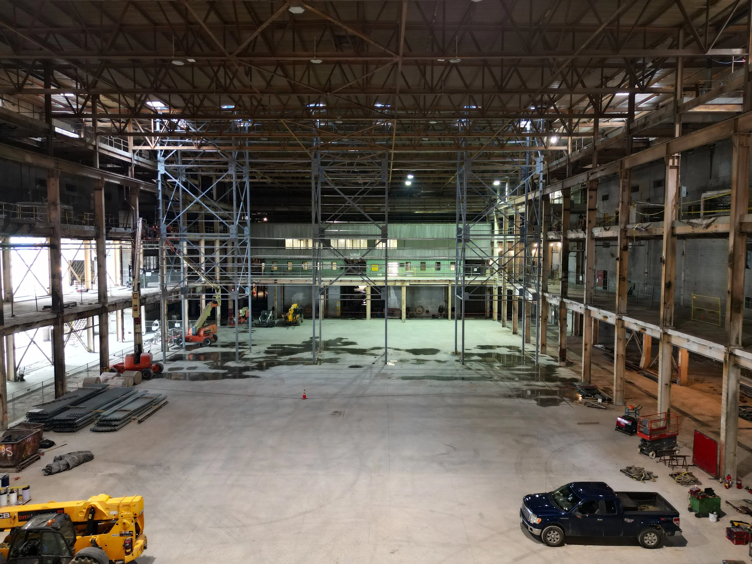 Interior of a large industrial or warehouse space under construction, with scaffolding, construction equipment, and vehicles visible.