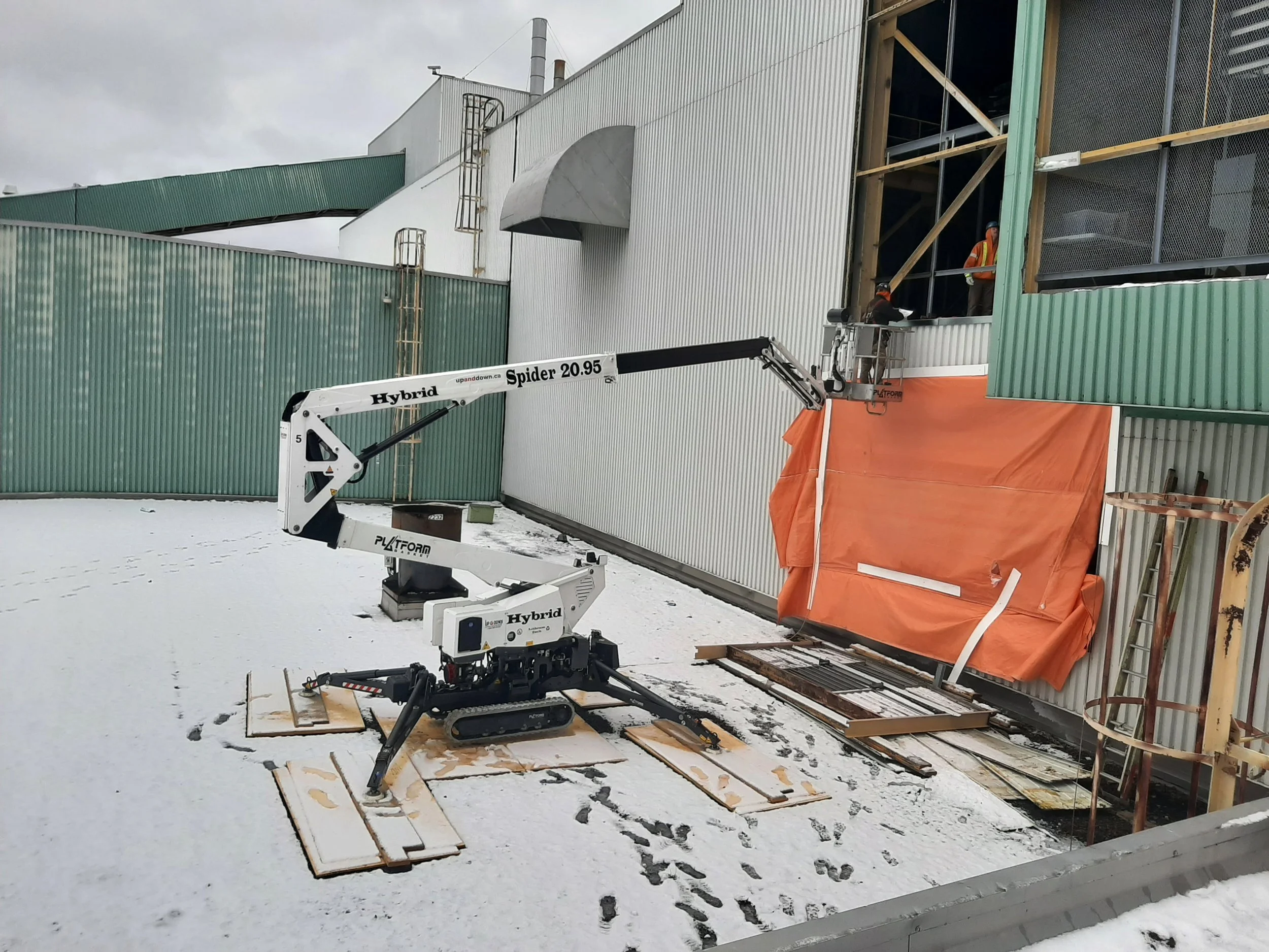 Construction workers using a boom lift to work on an upper window of a building with green and white exterior walls, snow on the ground.