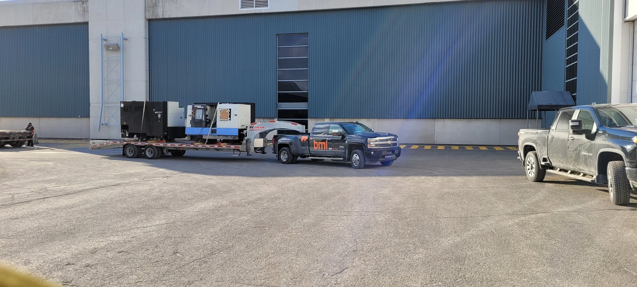 A vehicle towing large industrial equipment on a trailer parked in front of a modern building with blue metal siding and concrete walls, and a black pickup truck parked nearby.