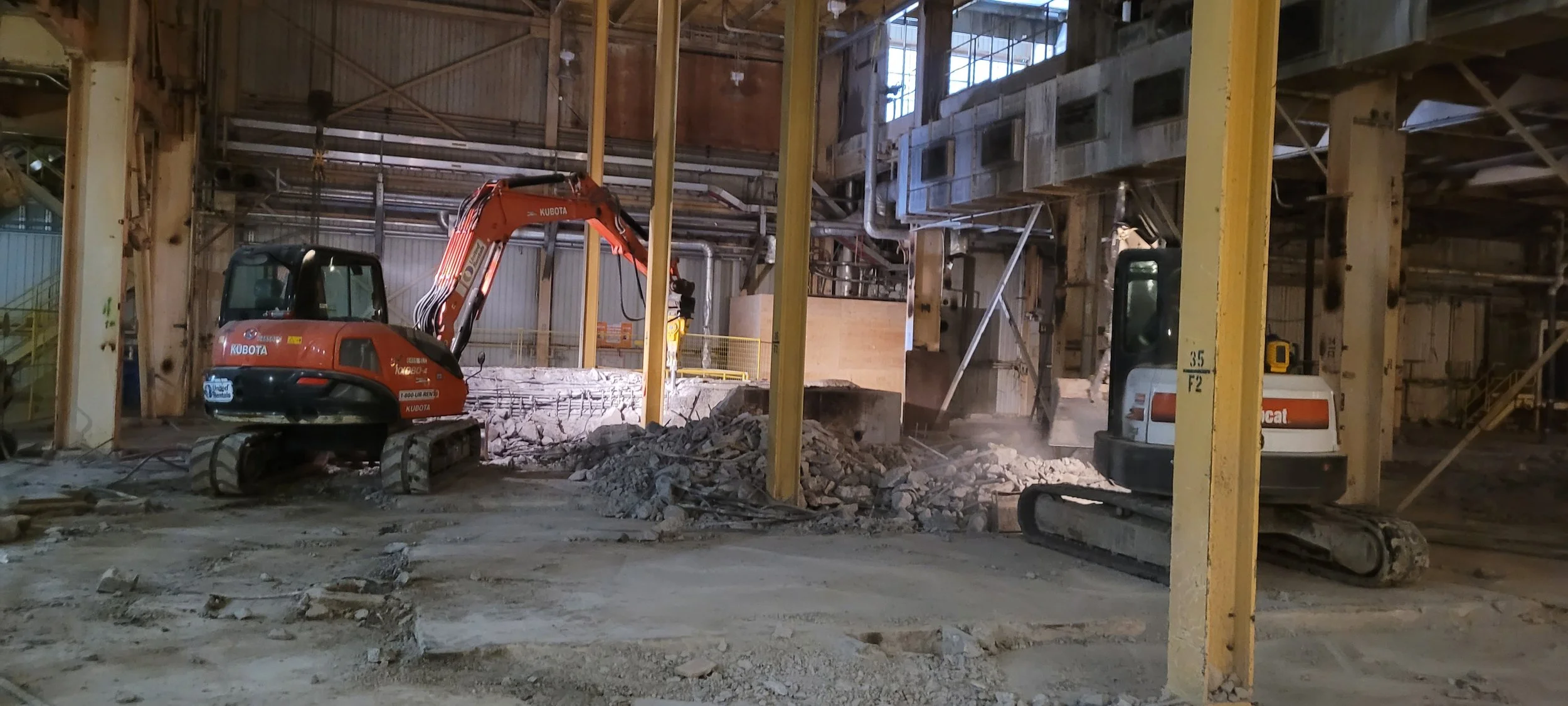 Construction site with two excavators working inside a large industrial building, surrounded by concrete debris and metal support beams.