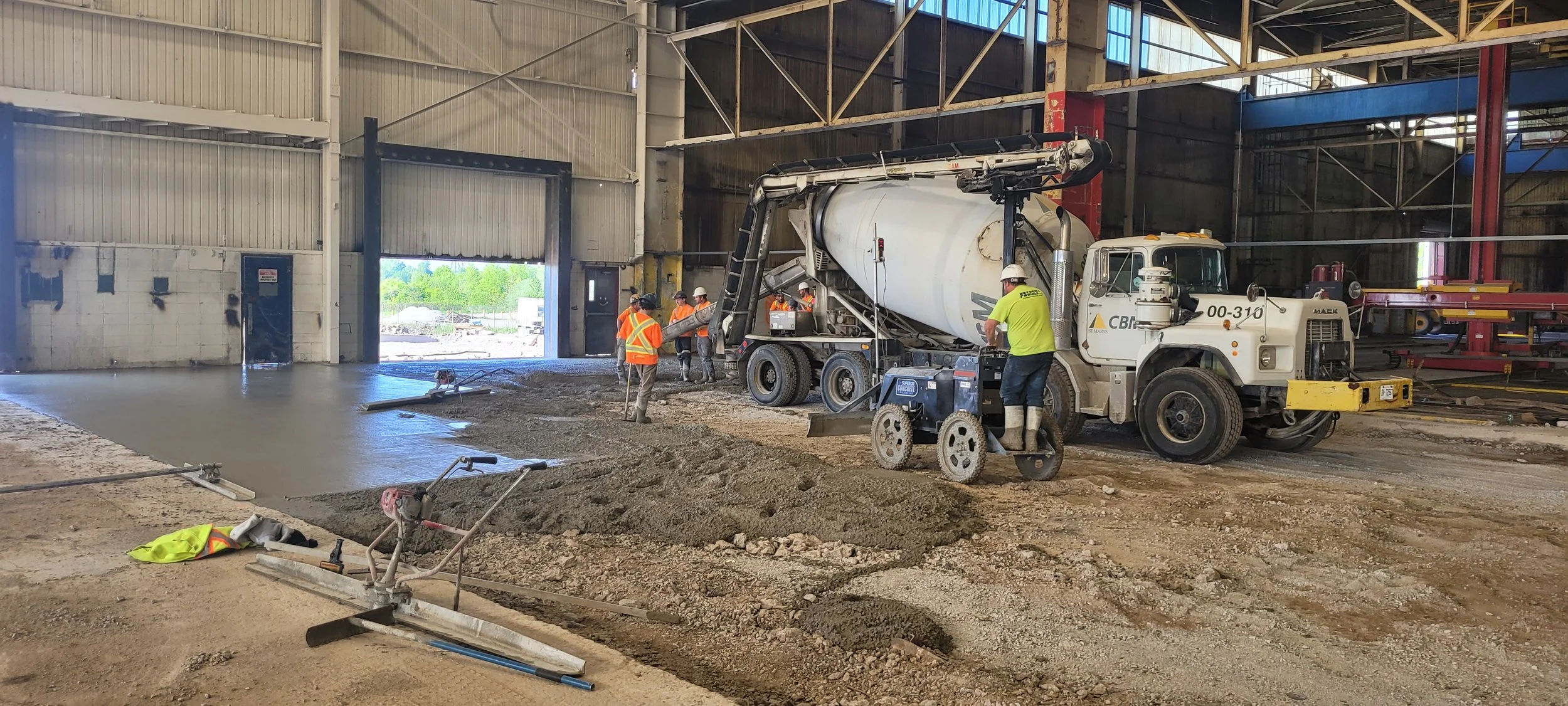 Construction workers are pouring concrete inside an industrial warehouse, with large machinery and equipment around.