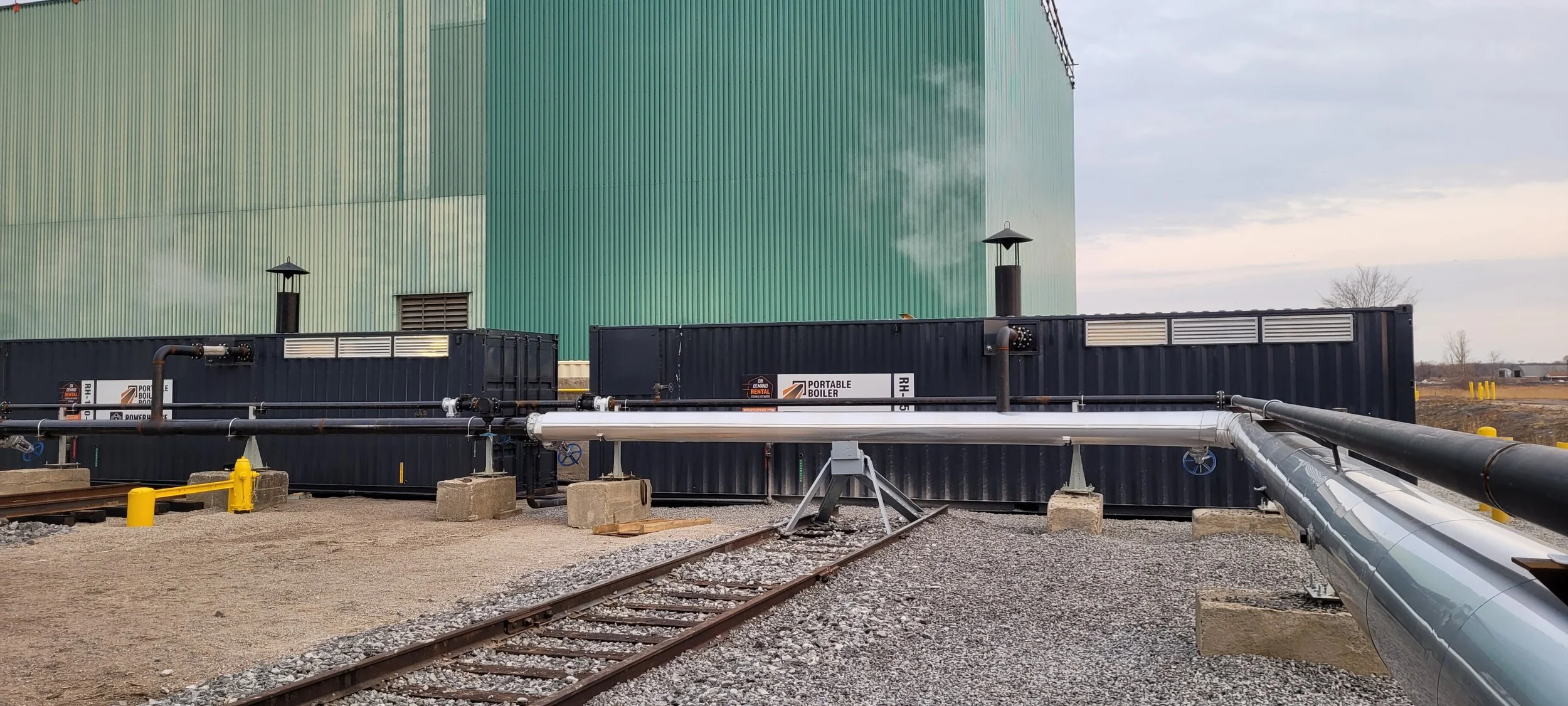Industrial area with a large green building, two black portable boilers, metal pipes, and railway tracks nearby.