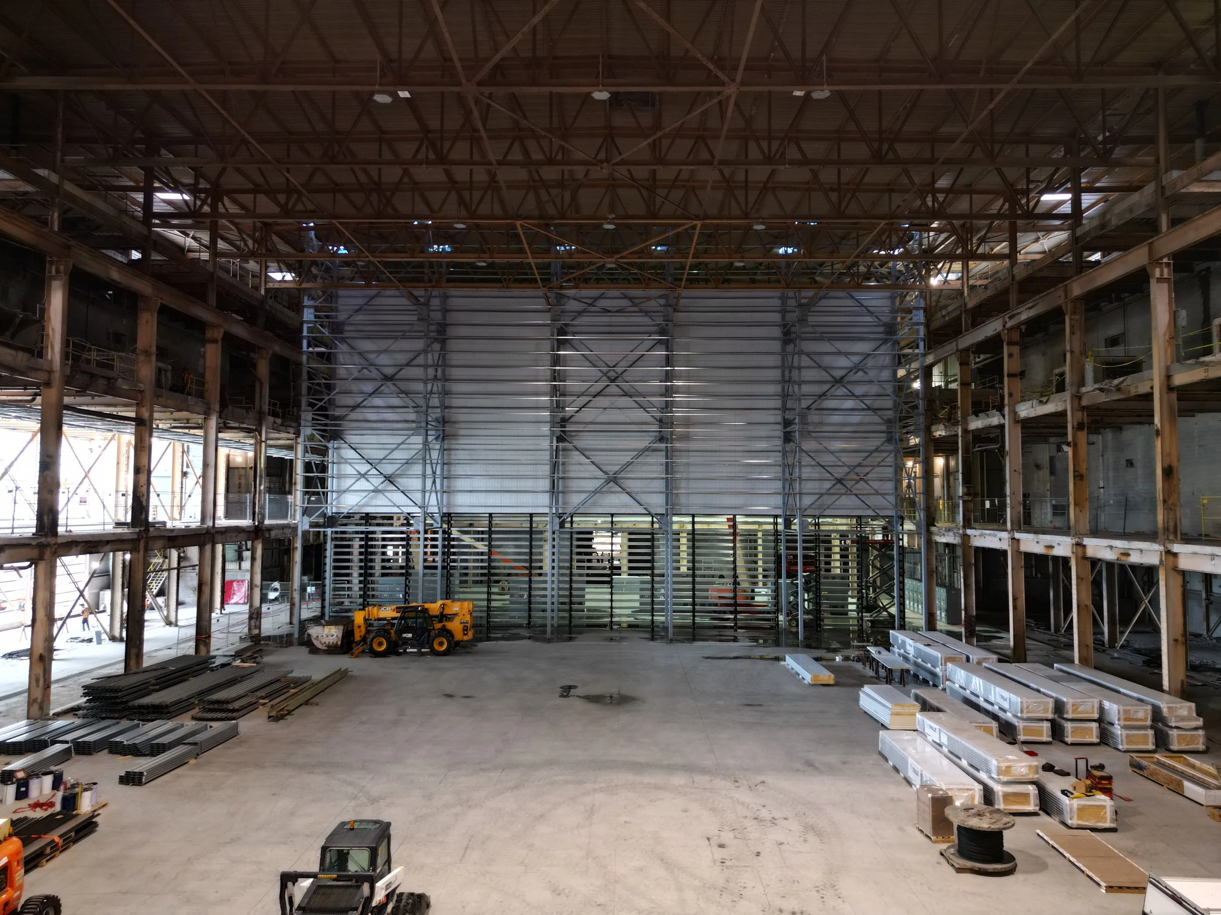 Construction site with interior steel framework, construction equipment, and prefabricated materials stored inside a large warehouse.