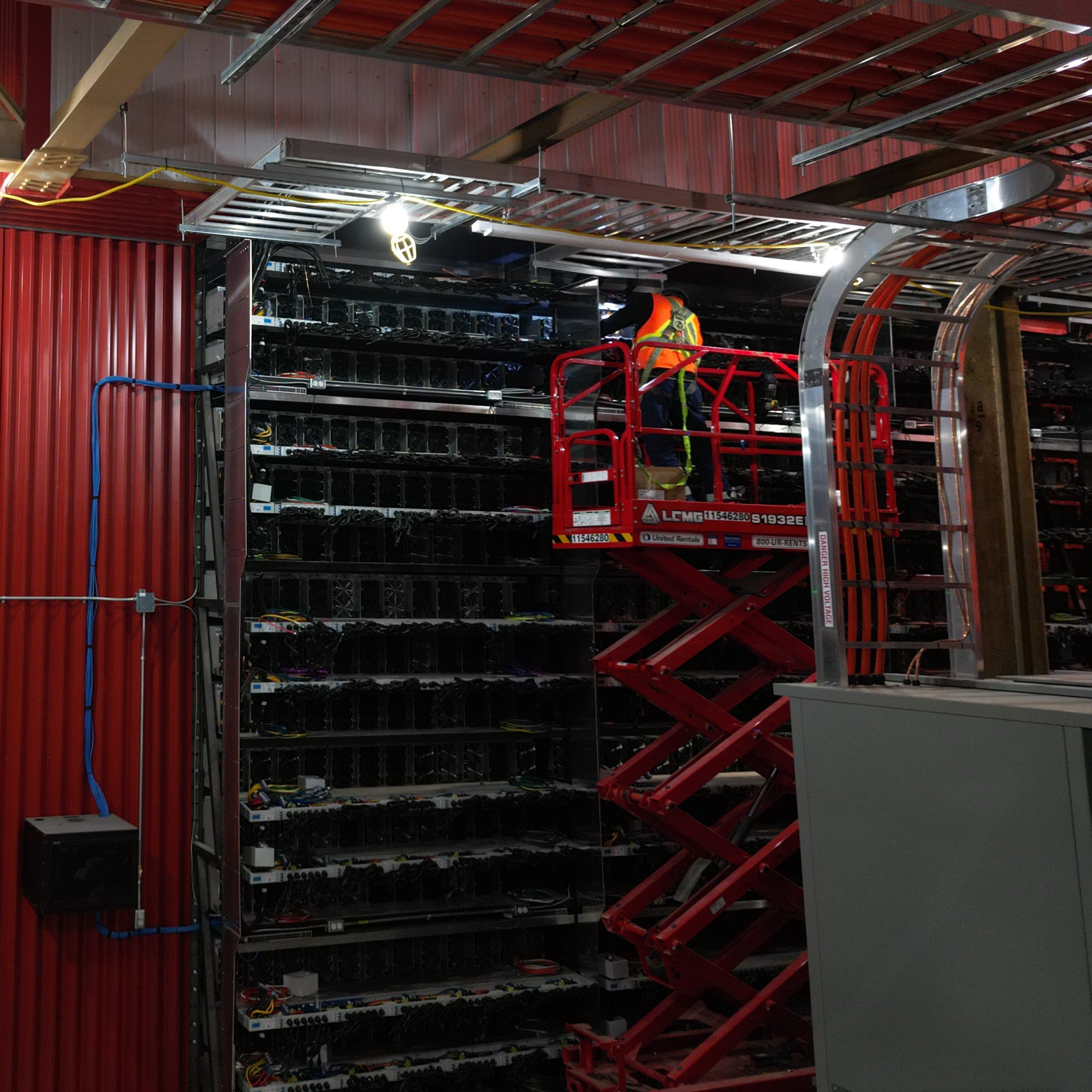 A worker in safety gear on a red elevated work platform working inside an industrial or data center with racks filled with electronic equipment and cables.