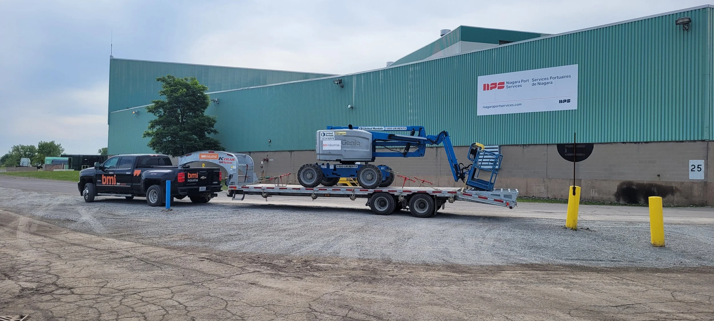 A black pickup truck with orange and gray branding is pulling a flatbed trailer carrying a blue cherry picker outside a large green building with signs indicating Niagara Port Services. The area has yellow and blue safety bollards and a gravel ground