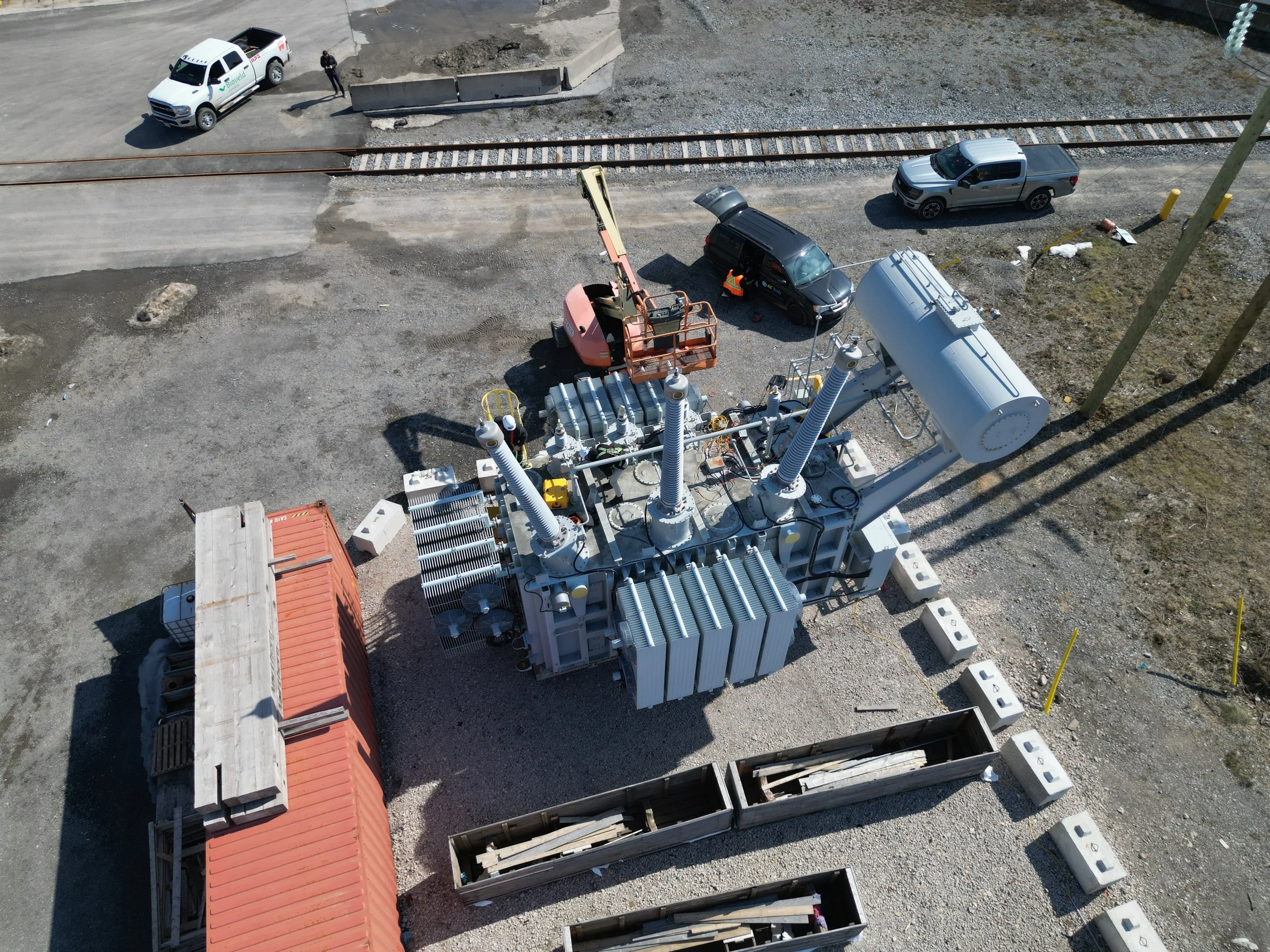 An aerial view of a construction site with electrical infrastructure, including large transformer and equipment, gravel ground, and several parked vehicles, including a police car, pickup trucks, and construction tools.