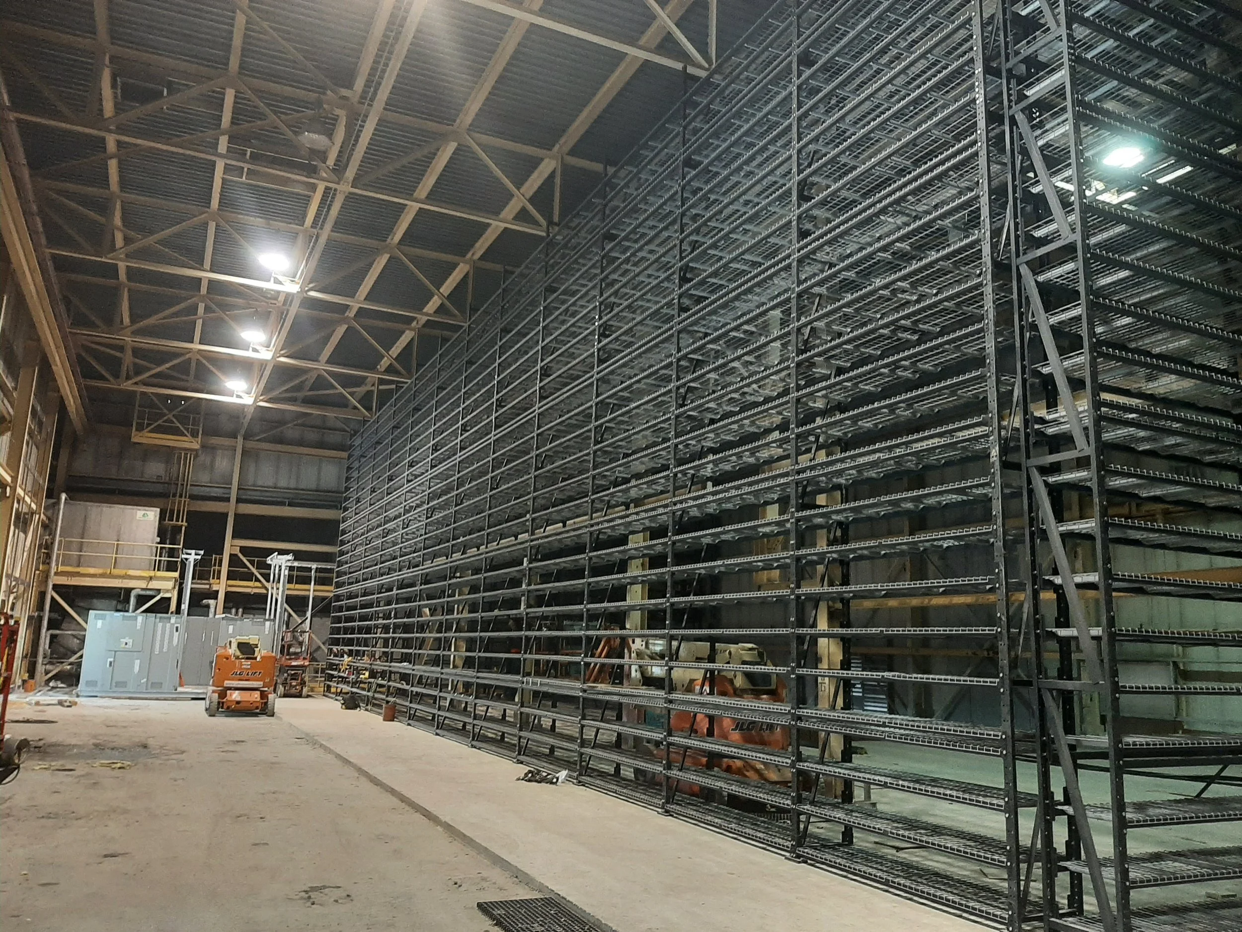 Interior view of a large industrial warehouse under construction with metal shelving units, construction equipment, and scaffolding.