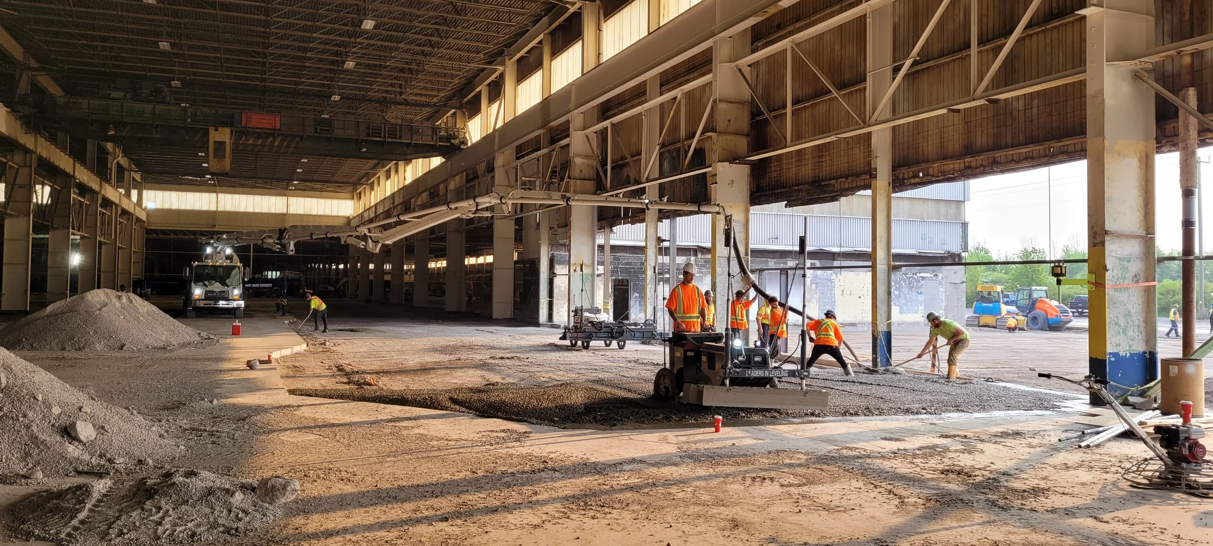 Construction workers in safety vests and helmets are working inside a large building under renovation, leveling and paving the ground with machinery, with construction equipment and vehicles visible outside.