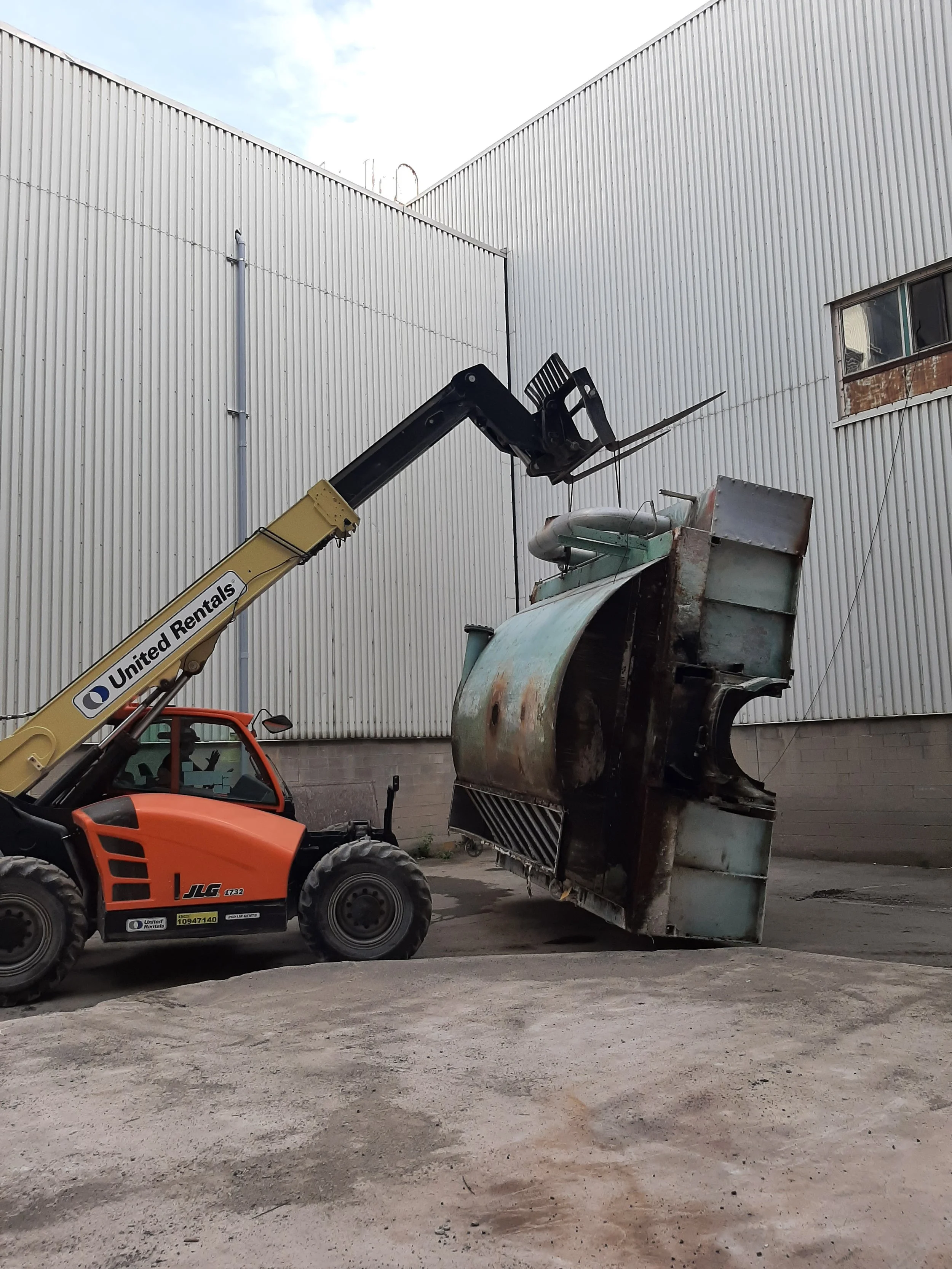 A forklift labeled 'United Rentals' lifting a large, rusted industrial machine outdoors between two large white industrial buildings.