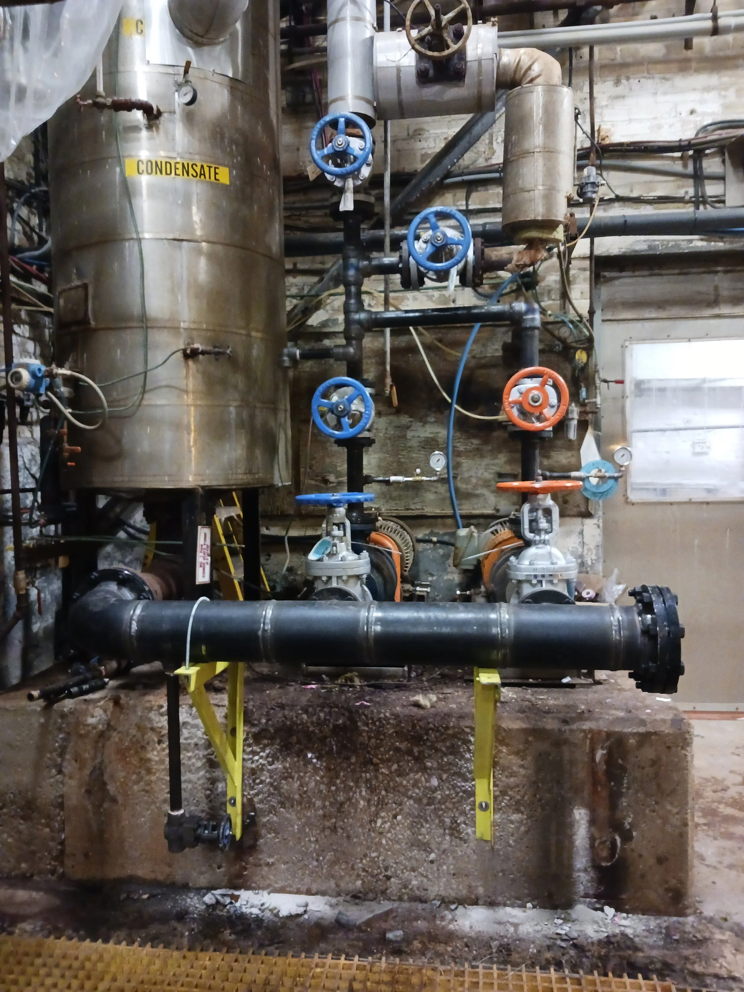 Industrial piping system with blue and orange valves, large metal tank labeled 'Condensate,' and gauges in an industrial setting.
