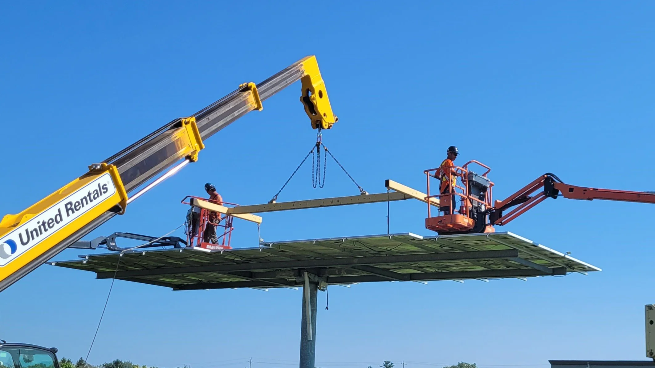 Two workers in safety gear are installing or maintaining an elevated billboard with solar panels, using a telescopic crane labeled 'United Rentals' on a clear blue sky day.
