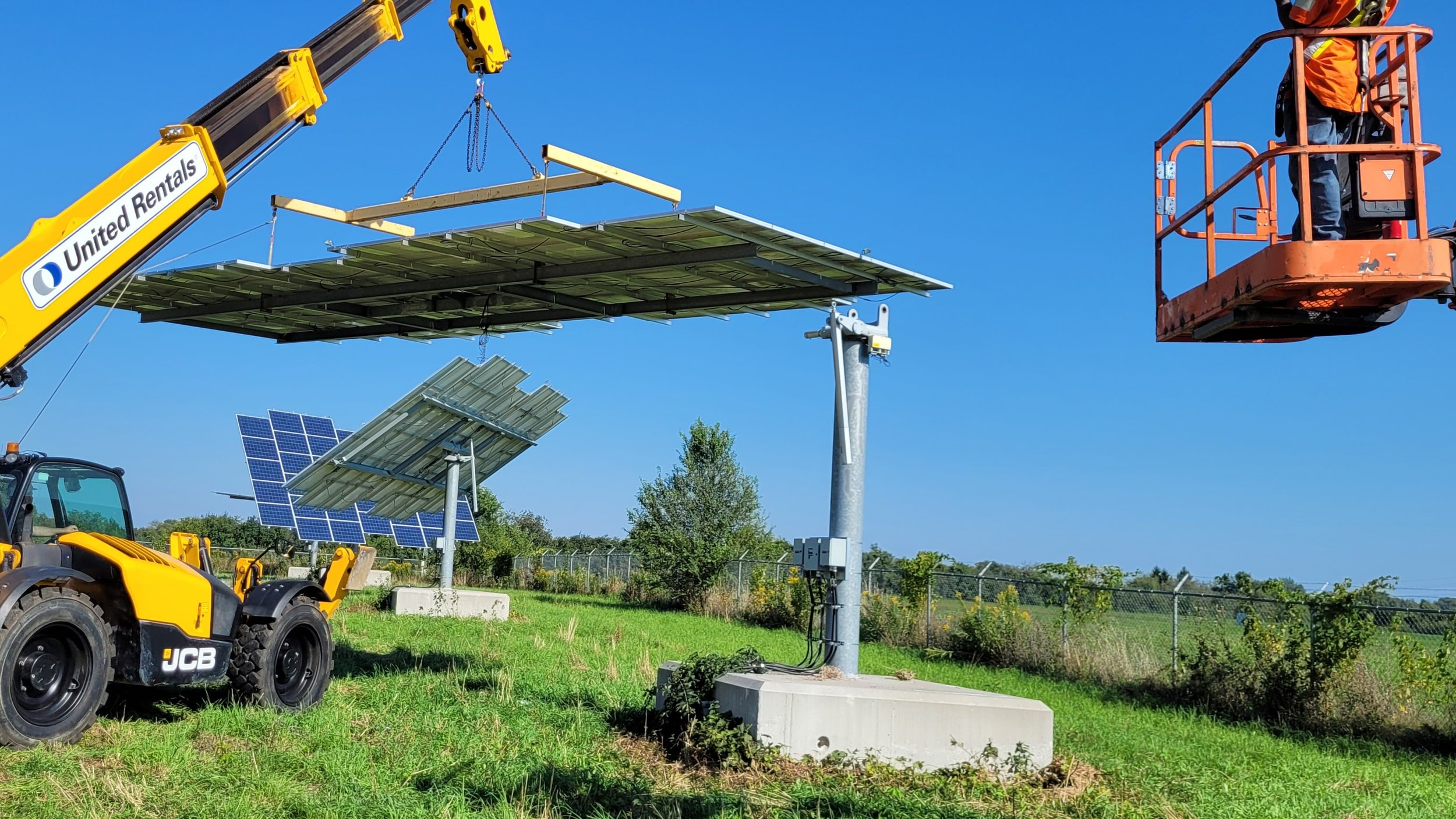 Solar panels being installed on a pole by construction workers using a crane and cherry picker at a grassy outdoor site.