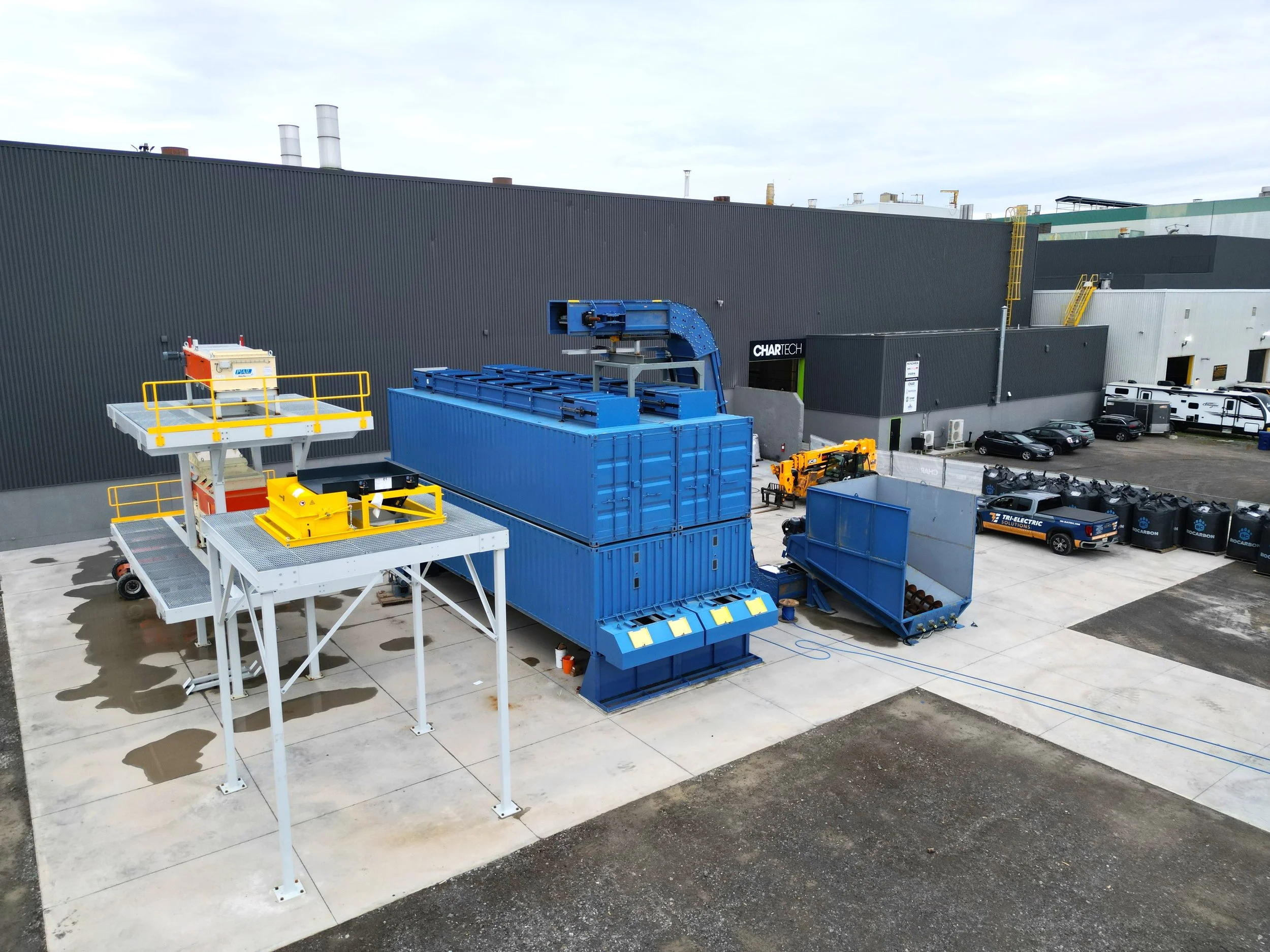 Industrial equipment and machinery outside a large warehouse building, including a blue container, yellow safety barriers, and a raised platform with railings, adjacent to a parking lot with several vehicles and RVs.
