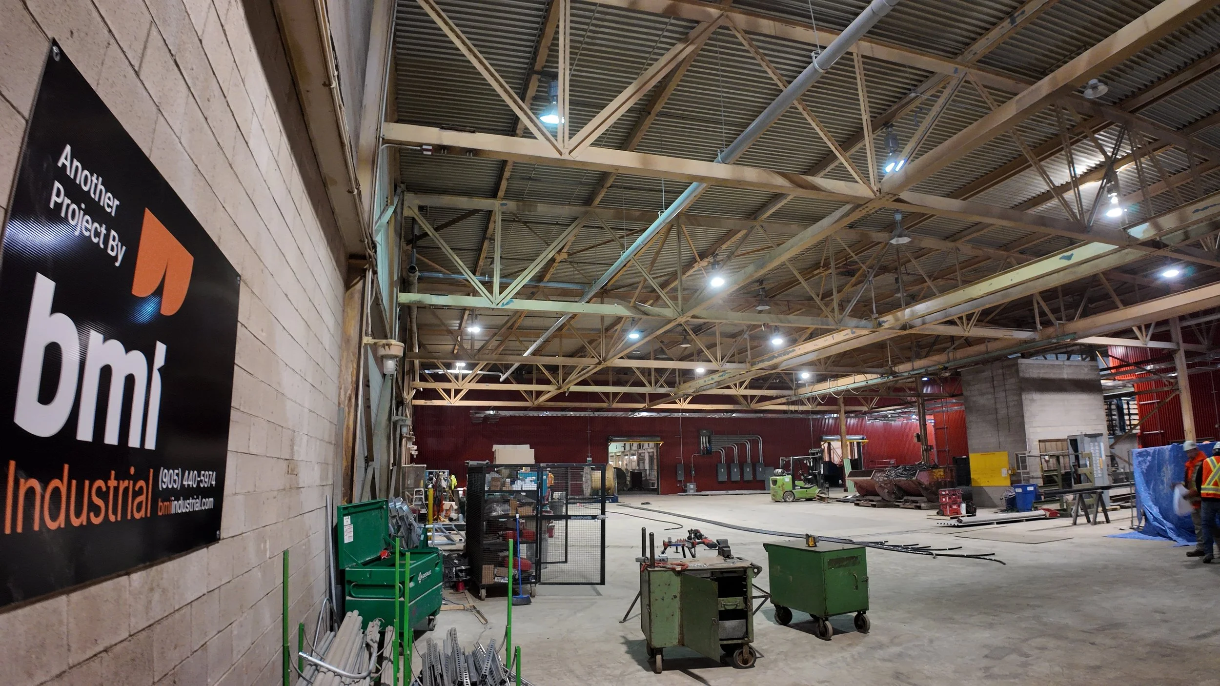 Interior of an industrial construction site with various equipment and workers. Large open space with high ceiling, metal trusses, and bright overhead lighting. Construction tools and materials are scattered on the floor.